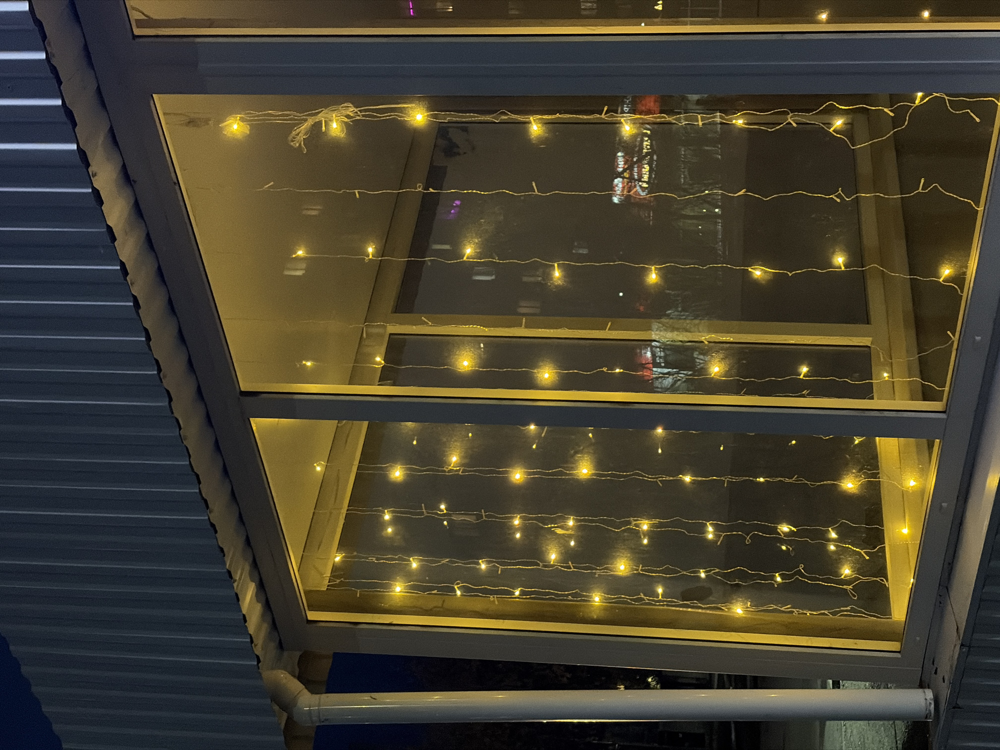 The image shows a rectangular skylight illuminated with strands of yellow fairy lights. The skylight is framed with a gray metal structure. The reflection in the skylight suggests a nighttime urban environment, with lights from distant buildings. The corrugated metal roof is visible to the left of the frame. A white pipe runs along the bottom of the skylight structure. No people are visible. There is no explicit reference to confirm that this is in Chaikovsky, Russia.