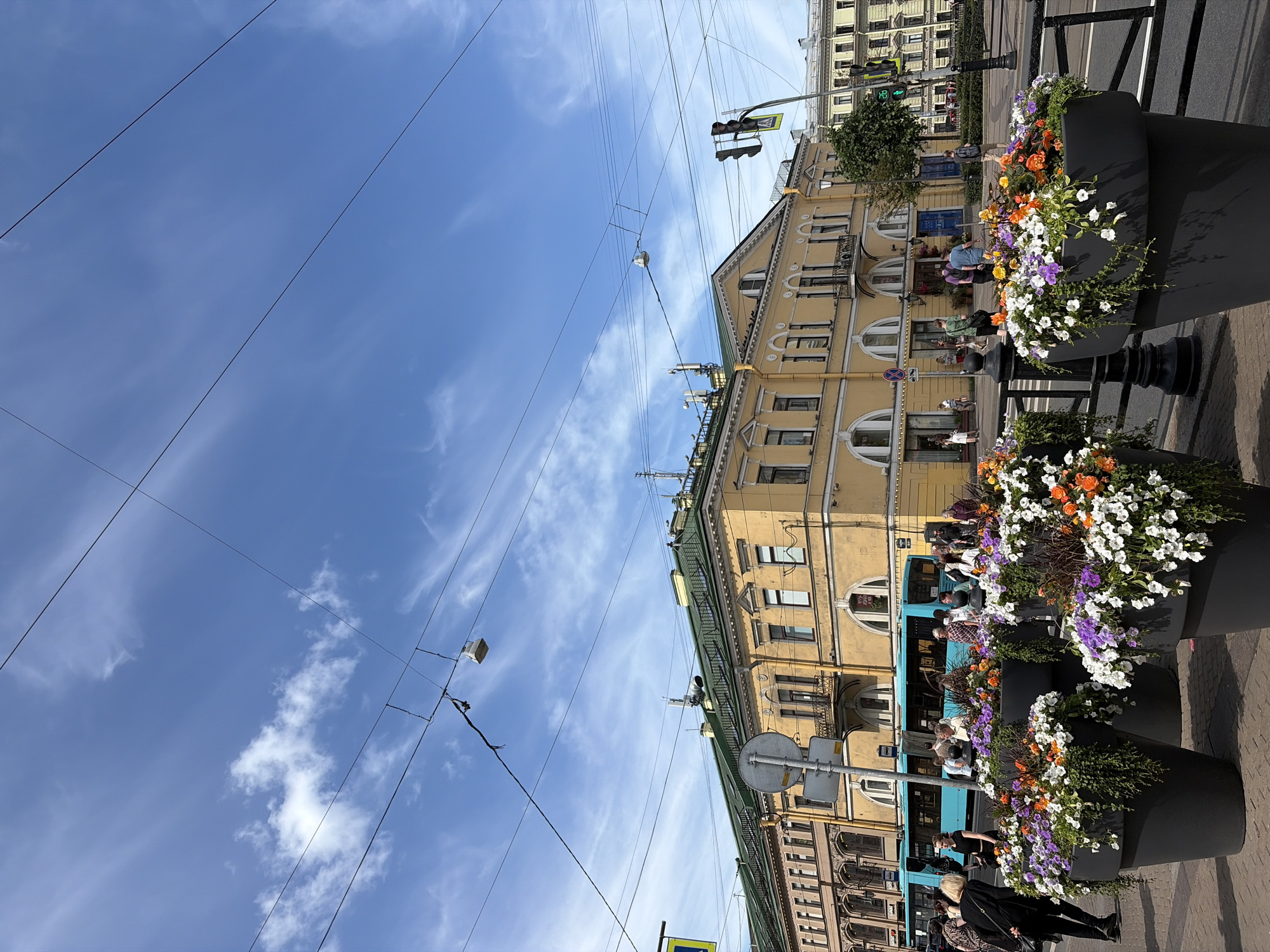 This image captures a street scene in Saint Petersburg, Russia, under a bright, partly cloudy sky. The perspective is from a low angle, looking up towards several buildings and a vast expanse of sky crisscrossed by numerous overhead wires.
The primary subjects are the buildings, which exhibit distinct architectural styles. The closest building is a tall, pale yellow structure with arched windows and decorative detailing. To its right, a teal-colored bus is partially visible, its windows reflecting the sky. In the foreground, large black planters overflowing with colorful flowers – white, purple, and orange – add a vibrant touch to the urban landscape.
Several people are scattered throughout the scene. Some are walking on the sidewalk near the buildings, while others appear to be passengers on the bus or are looking at the flowers. One woman in the foreground is walking away from the camera, dressed in black. The presence of a traffic light and road signs indicates an active street.
The time of day appears to be daytime, likely late morning or afternoon, given the bright sunlight casting shadows. The weather is pleasant, with a clear blue sky punctuated by wispy white clouds. The overall mood is one of a bustling city on a fair day, with the floral displays adding a touch of natural beauty to the urban environment.
No visible text is legible in the image. The architectural style of the buildings and the presence of overhead wires are characteristic of Saint Petersburg.