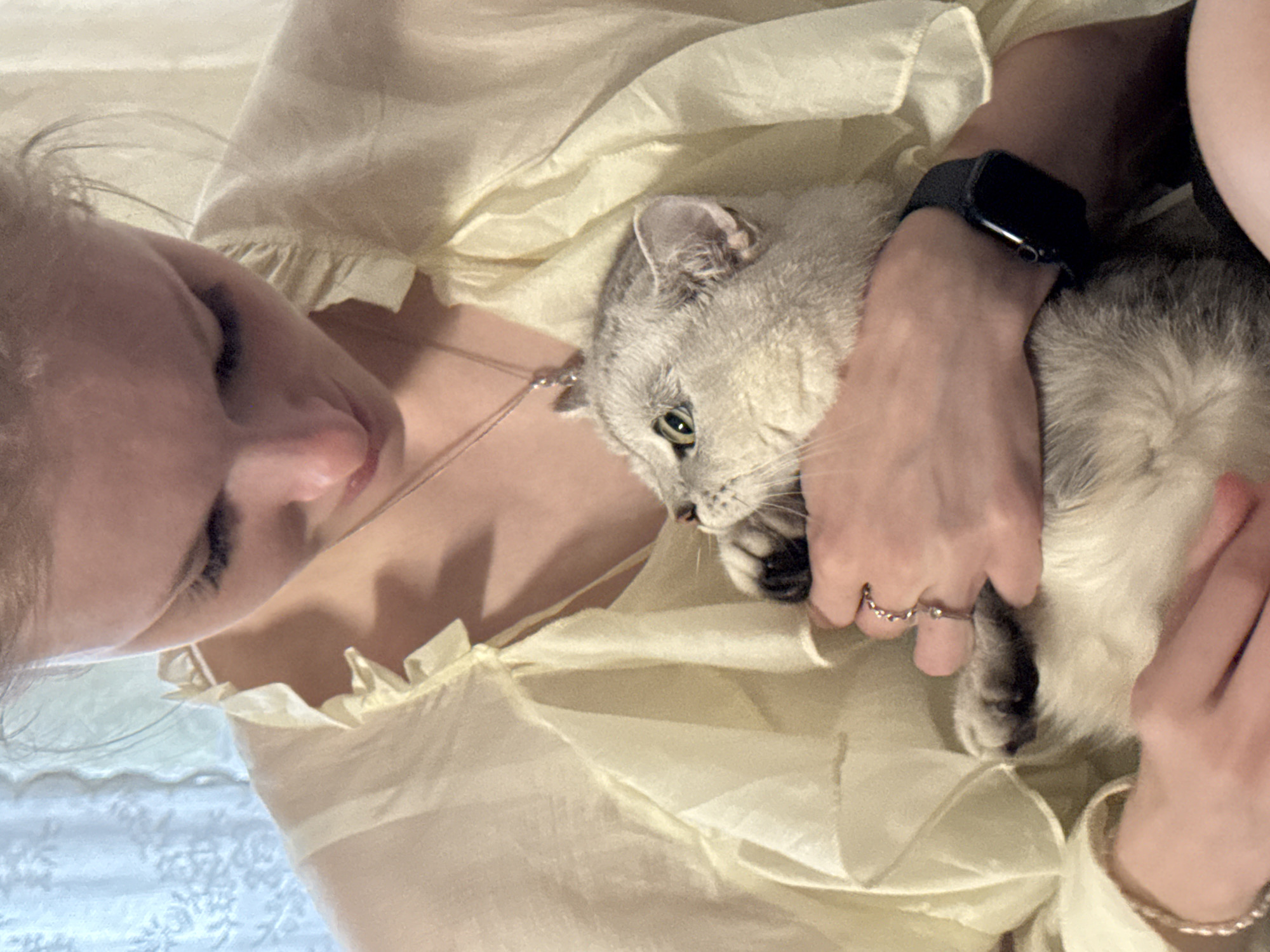 A person is holding a light gray cat close to their chest, their face is partially visible, looking down at the cat with a soft expression. The cat, with green eyes, is nestled in the person's arms and appears relaxed. Both subjects are wearing a light yellow, ruffled garment, possibly a dress or top. The person has a smartwatch on their left wrist and is wearing a ring on their right hand. The background is softly lit, suggesting an indoor setting with natural light. The overall scene evokes a feeling of tenderness and affection.