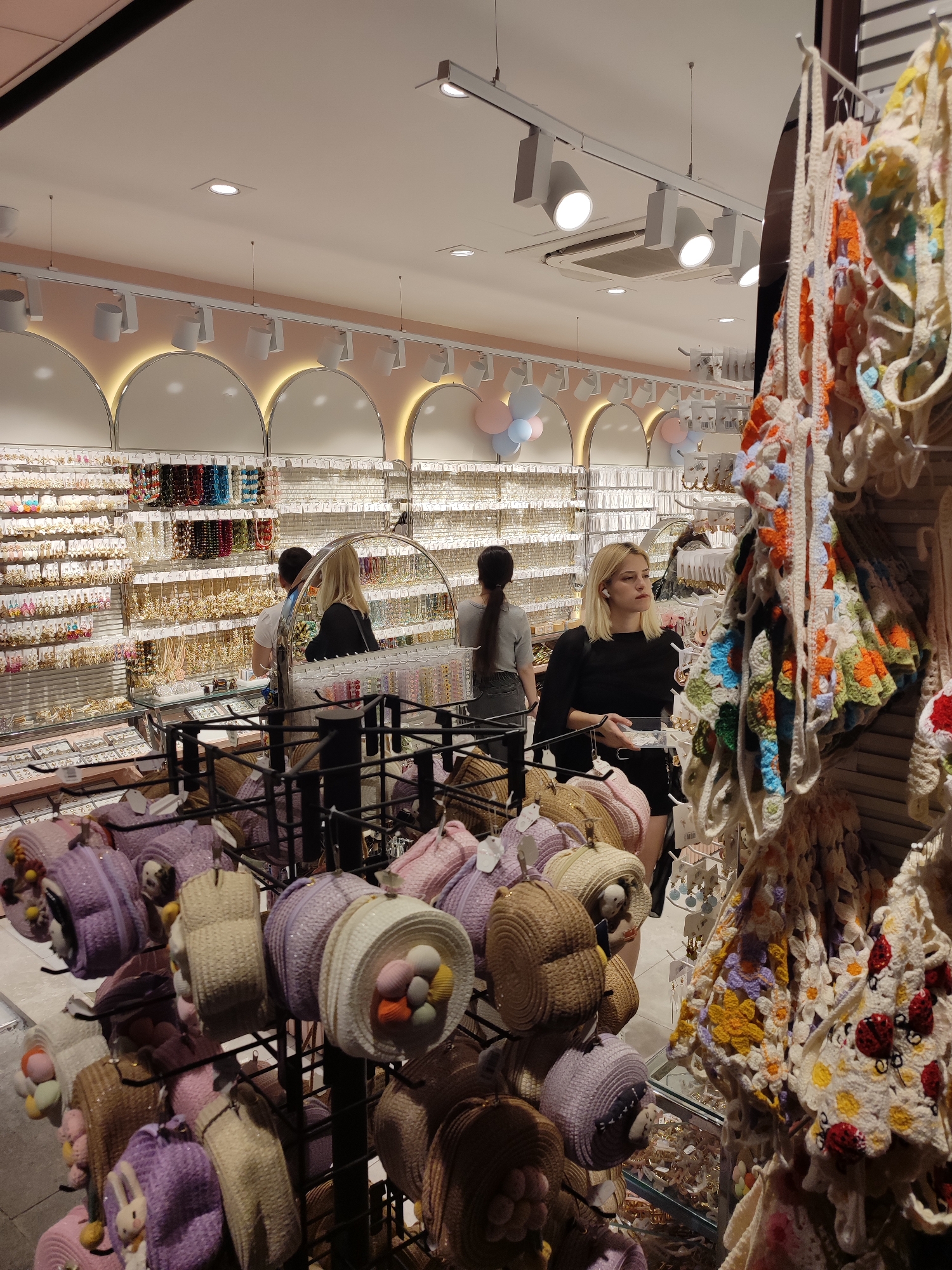 A bright, well-lit retail store interior is depicted, showcasing a variety of accessories. The store appears to be a boutique specializing in jewelry and possibly small bags or hats, with shelves and display racks filled with items. In the foreground, numerous straw hats or bags with colorful embellishments are displayed on black metal racks. Some have knitted or crocheted elements, with pom-poms and character designs such as rabbits. In the mid-ground, a blonde woman in a black top is visible, holding a clear plastic container and looking to her left. Behind her, a person with dark hair and a grey top is seen. The background reveals extensive displays of necklaces, earrings, and other jewelry items organized on white racks against a pastel pink wall. Arched architectural details and track lighting are prominent features of the store's ceiling and upper walls. The overall impression is that of a contemporary, fashion-oriented retail environment. This store is located in Kadıköy, Turkey, a district known for its vibrant shopping streets and boutiques.