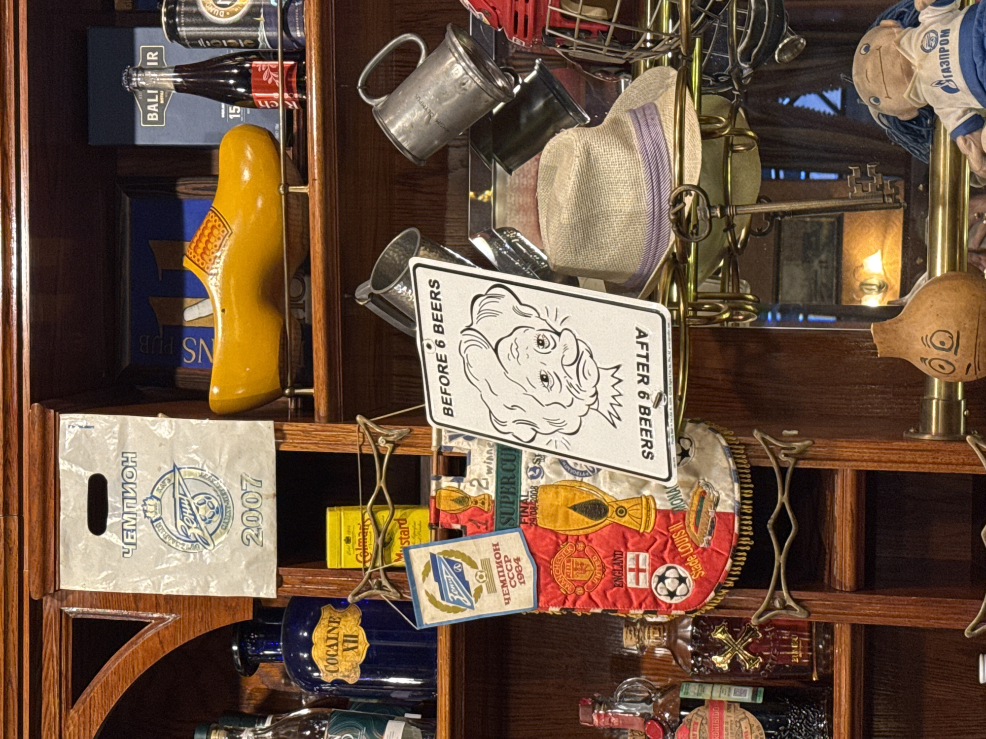 The image depicts a shelf filled with various items, likely in a bar or establishment in Saint Petersburg, Russia, given the presence of Cyrillic text. On the top shelf, a white paper bag with "ЧЕМПИОН" (Champion) and "ЗЕНИТ" (Zenit) logos, along with "2007," is displayed. Next to it, a yellow wooden clog and a bottle of Balisir.
Below, a dark blue bottle labeled "COCAINE XII" is visible, alongside a yellow tin of Colman's Mustard. A red and white pennant with the Manchester United logo and "ENGLAND" is hung, next to a trophy graphic that reads "SUPER CUP FINAL 29/08/2008." A sign with a cartoon face and the text "BEFORE 6 BEERS" and "AFTER 6 BEERS" is also present.
On the right, several metal mugs are placed, one with "OLYMPUS" inscribed. A straw hat sits below these mugs. Further down, glass bottles with red and gold accents are visible, one with a label that appears to have a prohibition symbol. A large ornate key hangs in front of a mirrored surface reflecting an interior with lighting. At the bottom, a gourd with painted eyes and a stuffed doll wearing a blue and white uniform with the Gazprom logo are present. The overall environment suggests a display of memorabilia and decorative items.