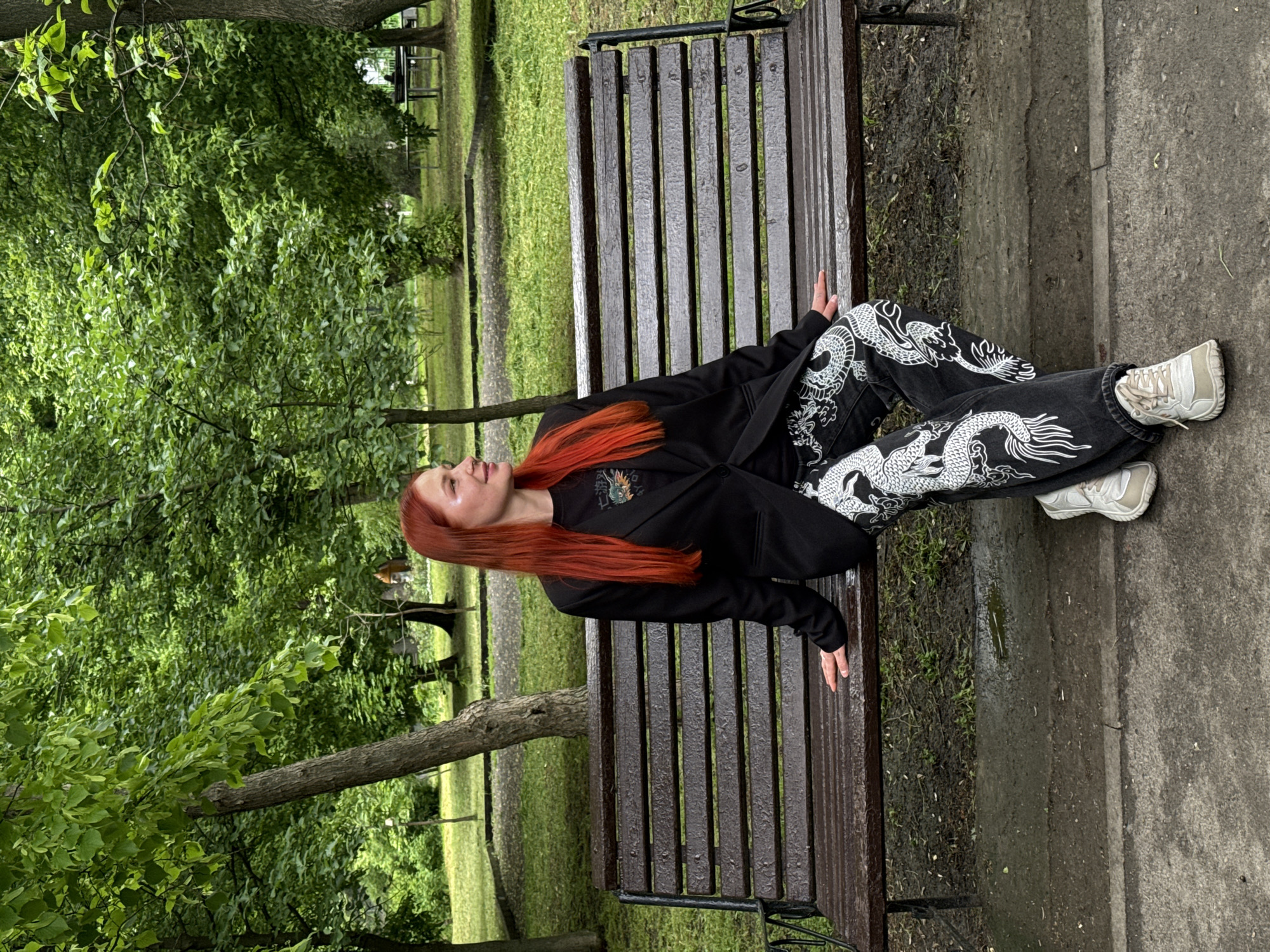 A young woman with vibrant red hair is reclining on a weathered wooden park bench. She is wearing a black jacket over a black t-shirt and striking black jeans adorned with white dragon designs. Her feet are clad in white sneakers. The bench is located in a park in Rostov-on-Don, Russia. Lush green trees and grass surround the bench. A paved pathway borders the bench, with patches of moss and grass visible between the stones.