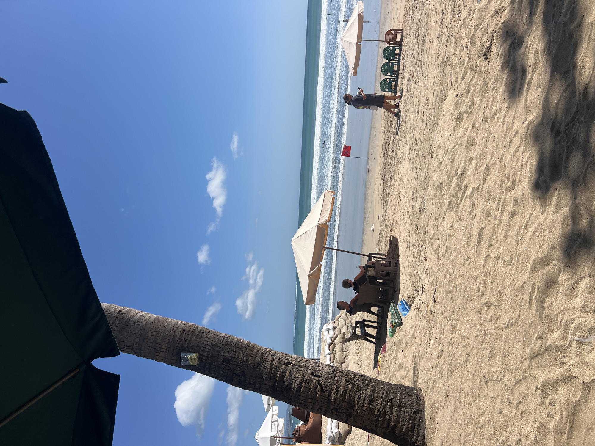 A tropical beach scene unfolds under a bright blue sky with scattered clouds. In the foreground, the coarse sand of Kuta Beach, Bali, Indonesia, is textured by ripples and shadows cast by unseen foliage. A thick, weathered palm tree trunk stands prominently on the left, its bark rough and fissured. A glass with a yellow liquid and ice is attached to the tree trunk. A large, dark green umbrella shades the immediate foreground.
Further back, a beige beach umbrella covers two individuals seated in chairs, facing the ocean. Beside them, colorful beach mats and bags lie on the sand. To the right, a woman wearing a dark top and shorts walks along the shore, holding a smartphone. She is observed by a group of green plastic chairs and one orange chair situated near another beige umbrella. A small red flag planted in the sand marks the shoreline. The ocean, with its gentle waves, extends to the horizon, where faint outlines of swimmers can be discerned. In the background, other beach umbrellas and beanbag chairs suggest a well-visited tourist spot.