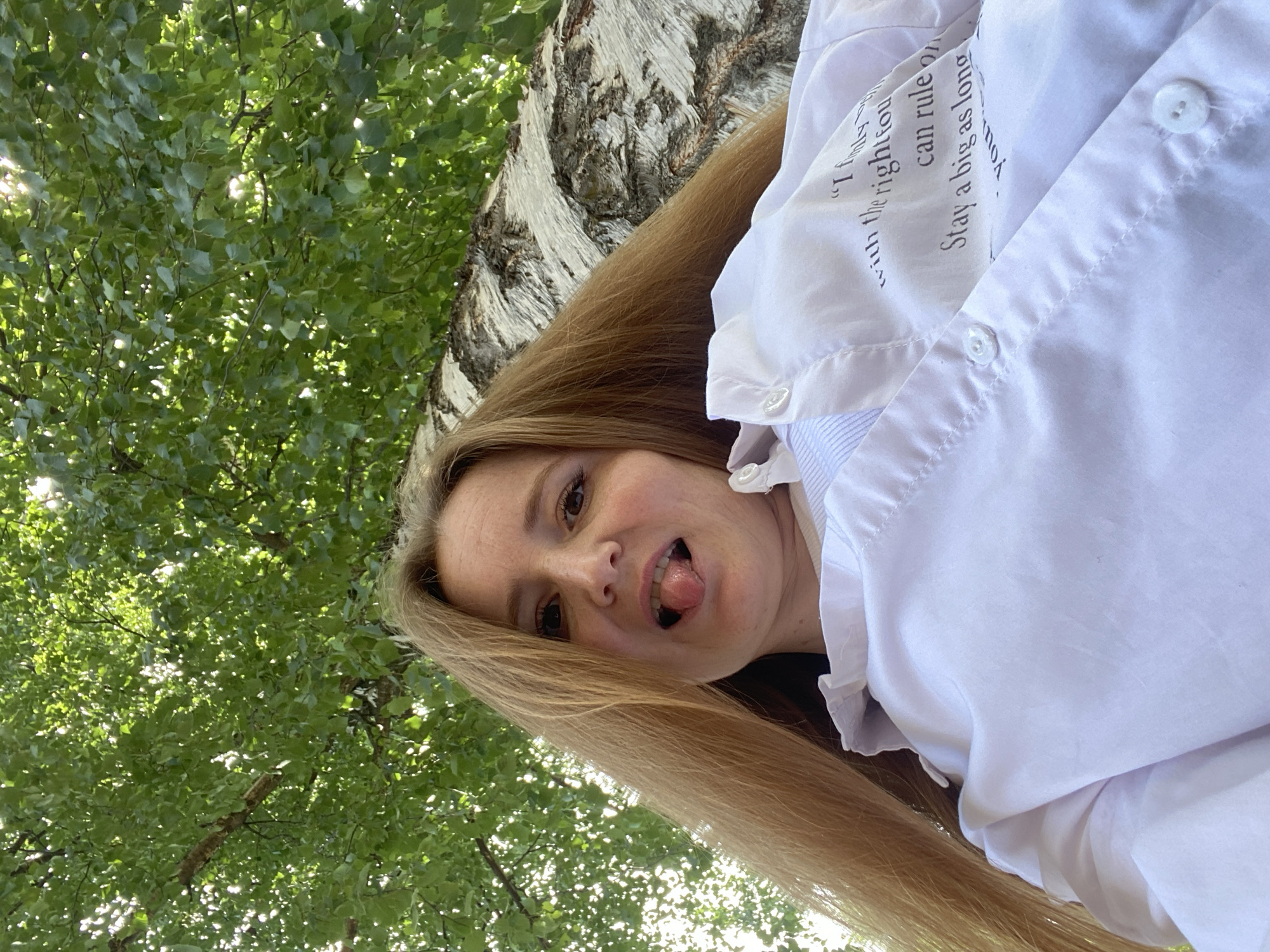 A low-angle, medium shot captures a young Caucasian woman with long blonde hair sticking her tongue out at the camera. She is wearing a white collared shirt with blue pinstriped fabric visible underneath at the collar and placket. The shirt has a ruffled detail along the collar. A light-colored, textured tree trunk, possibly birch, is visible behind and to the right of her. Lush green leaves and branches of a tree canopy are overhead, dappled with sunlight. The scene appears to be outdoors in a park or natural area within the city of Chaikovsky, Russia.