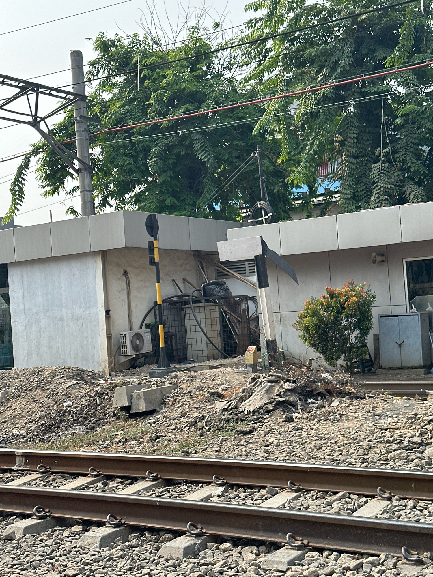 The image depicts a view of railway tracks with ballast and concrete sleepers in the foreground. In the background, a low-rise building with a grey facade and grey paneling on top is visible, surrounded by trees and vegetation. Adjacent to the building, there are utility poles, overhead electrical wires for the railway, an air conditioning unit mounted on the side of a concrete structure, a traffic signal with black and yellow stripes, a metal cage containing some items, debris, and a small evergreen bush. There are no people visible in the image. The overall scene suggests an outdoor, urban or semi-urban environment, possibly near a railway station or maintenance area.