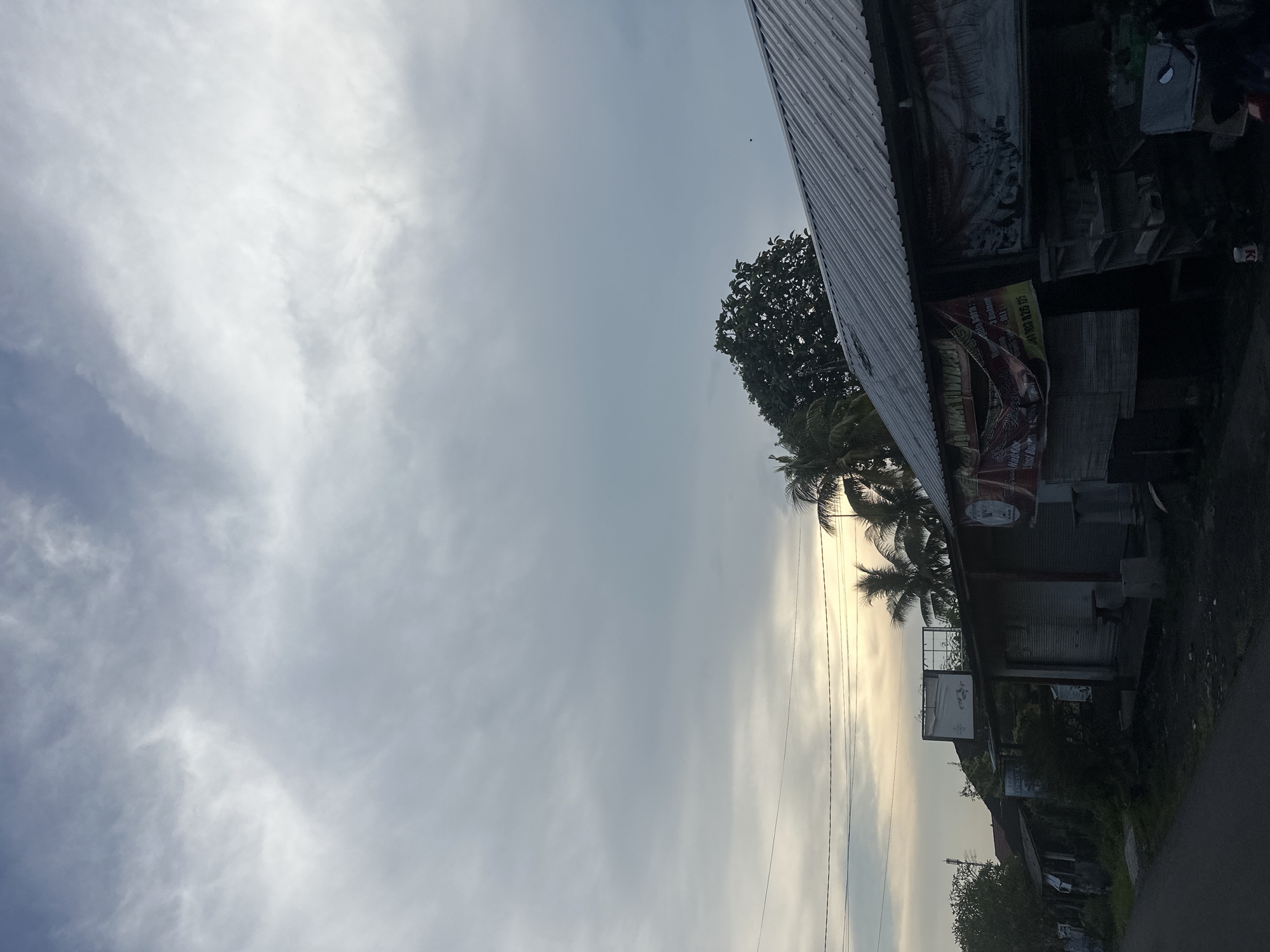 The image presents a partially obscured view of what appears to be a street scene in Kekeran, Indonesia, at or around sunrise or sunset given the position of the sun behind the palm trees, with a cloudy sky. The perspective is from the side of the road. A building with a corrugated metal roof dominates the right side of the frame. Signage is visible on the building's façade, but the text is partially obscured and illegible. Palm trees are present, silhouetted against the sun. Power lines are visible. Vegetation is present along the roadside.