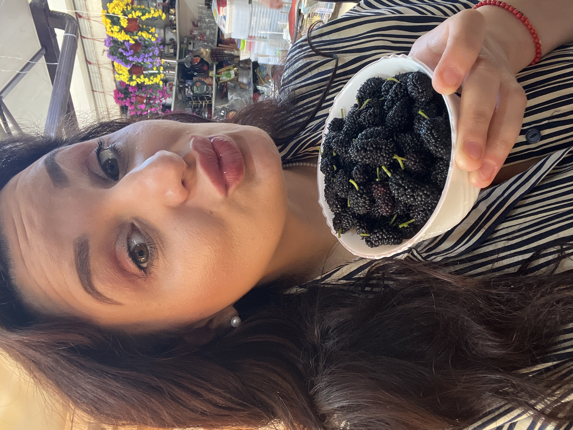The image displays an adult female in an indoor retail or storage environment, location Unknown, Unknown. She is depicted from the upper torso, facing the camera with her lips pursed in a kissing gesture. Her eyes are open, with visible eye makeup including eyeshadow, eyeliner, and mascara. She possesses long, dark brown, wavy hair. A light-colored stud earring is present on her left ear, and a pearl necklace is partially visible around her neck. She wears a horizontally striped shirt, featuring dark and light bands, and a red beaded bracelet on her right wrist.
In her right hand, she holds a white, scalloped-edge ceramic bowl filled with numerous dark purple-black mulberries. Short, green stems are discernible on many of the mulberries.
The background, out of focus, contains shelving units stocked with various household goods and plastic containers. Above the shelves, artificial flower arrangements in pink, purple, and yellow hang. A white ceiling with a visible dark pipe or gutter system extends horizontally across the upper frame.