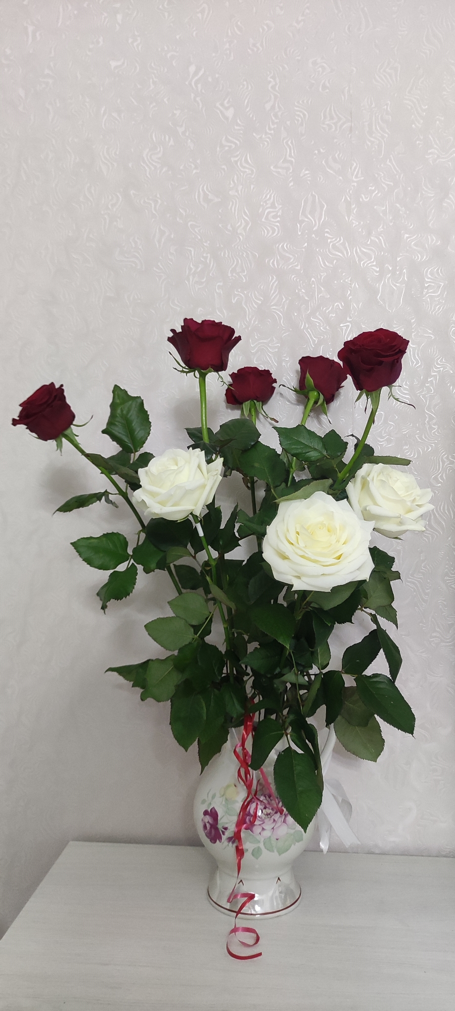 A vase of red and white roses sits on a table against a patterned off-white wall. The vase is cream-colored with a floral design in purples and greens. Six red roses and four white roses are visible. Red and white ribbons are around the neck of the vase. The scene is located in the city of Chaikovsky, Russia.