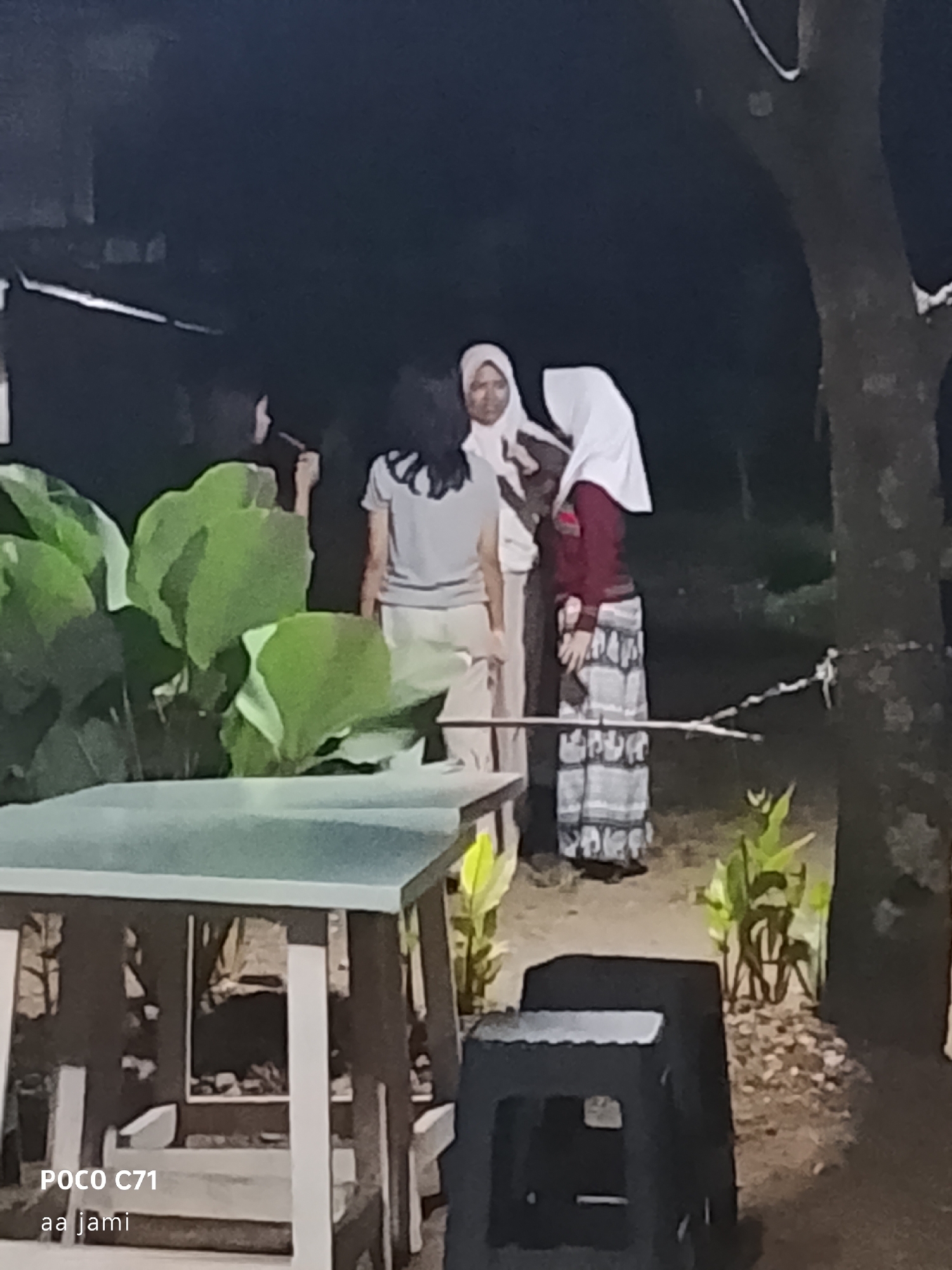 The image shows a group of four individuals standing outdoors at night. In the foreground, a table and a black stool are visible. The background features large green leaves, a tree trunk, and foliage. One person is wearing a white headscarf and a maroon top with a patterned skirt. Another person wears a white headscarf and a light-colored shirt and pants. A third individual is wearing a gray t-shirt and light-colored pants, and a fourth person's attire is not clearly discernible. The scene suggests an informal gathering or interaction. The presence of the text "POCO C71" and "aa jami" in the lower left corner suggests the image was taken with a Poco C71 mobile phone and may be related to a person or group named "aa jami." Without further information, it is impossible to pinpoint the exact location within Tasikmalaya, Indonesia.