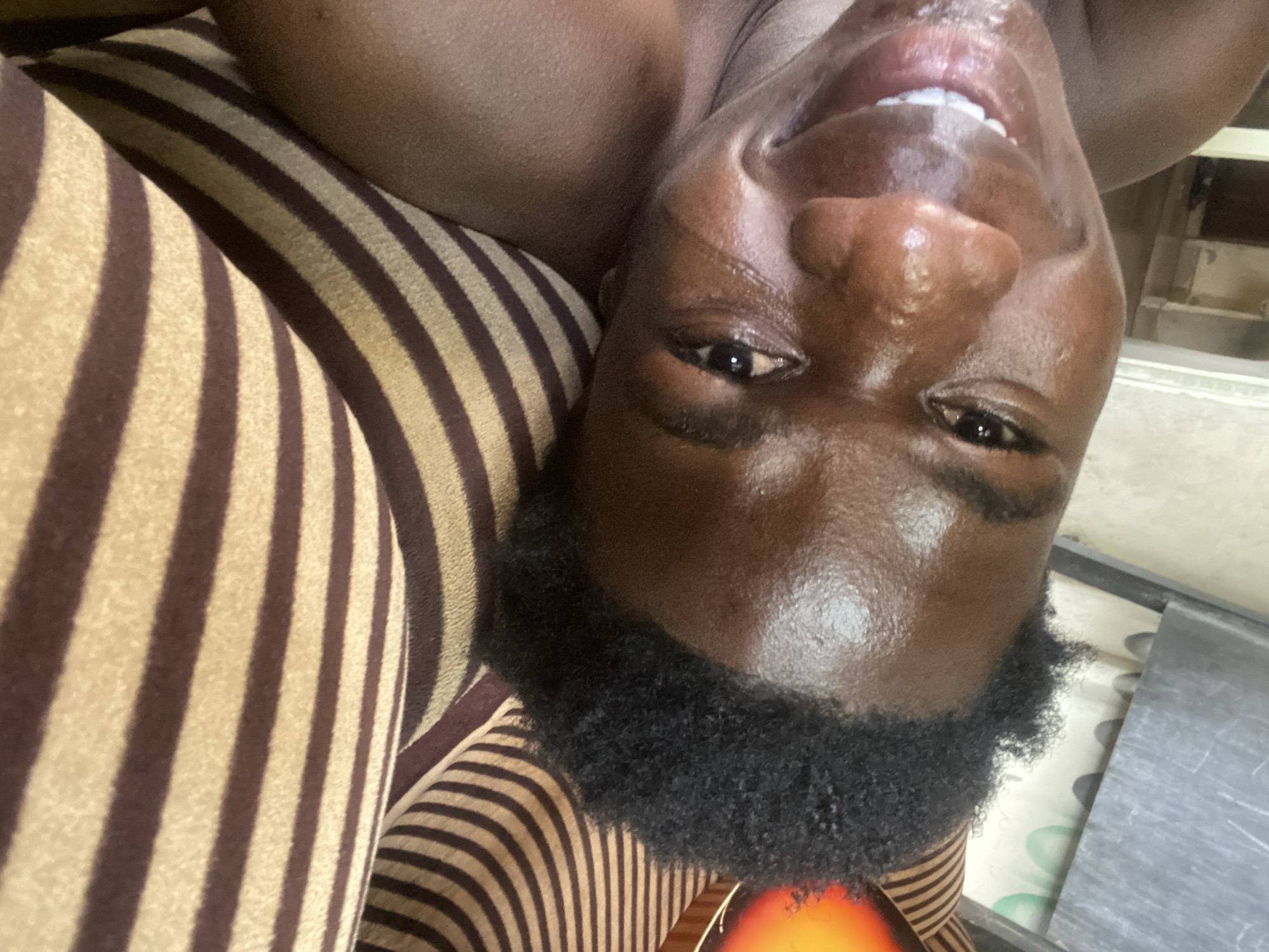An upside-down, close-up selfie of a dark-skinned person with short, dark, curly hair, appearing to smile. The person's face is shiny. They are leaning against a surface with vertical brown and tan stripes. There is a glimpse of a table/surface with a silver colored top visible to the right of the person's head. There is not enough information in the image to confirm the location.