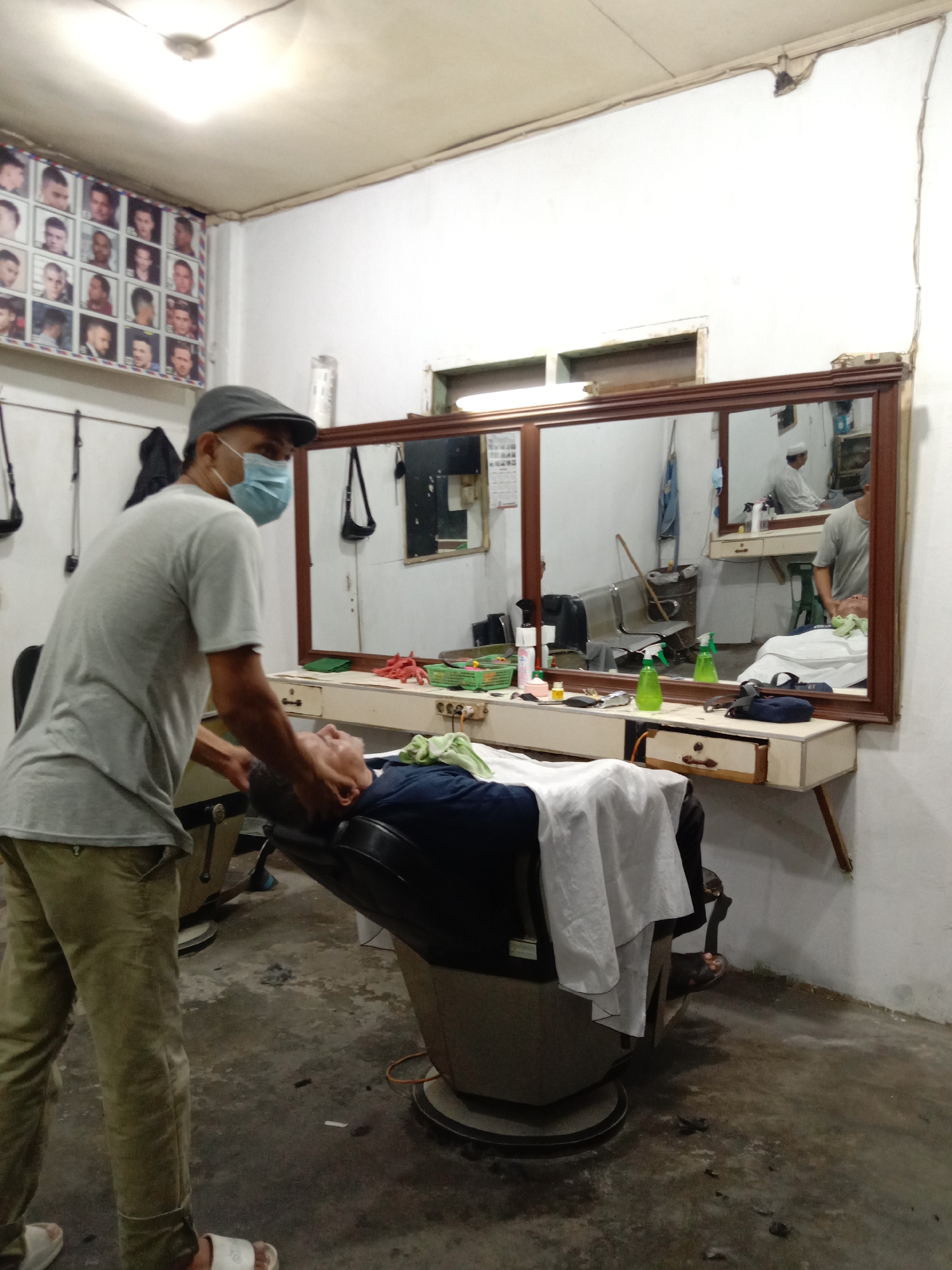 A man wearing a grey t-shirt, khaki pants, a grey cap, and a light blue face mask is standing next to a barber chair where a man is lying down. The man in the t-shirt appears to be attending to the man in the chair. The scene takes place in what appears to be a barber shop. A large mirror hangs on the wall behind the chair. Below the mirror, there is a counter with various barber tools, including green spray bottles, combs, and containers. On the left wall, there is a poster displaying various men's hairstyles. The floor is stained and has hair clippings scattered across it. This is possibly in Lhokseumawe, Indonesia.