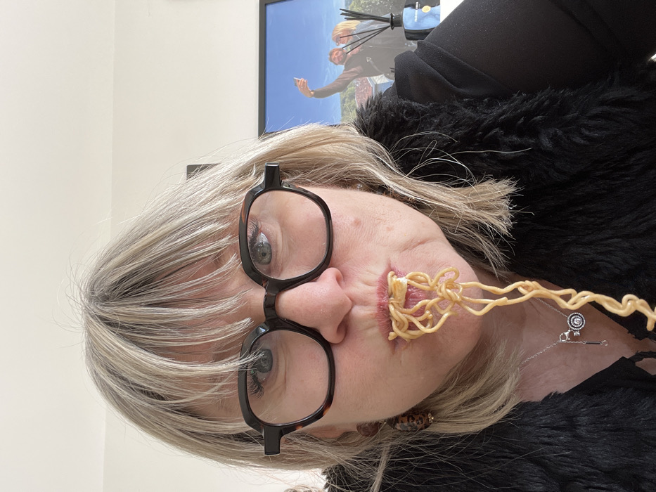 A frontal, close-up image depicts a middle-aged female subject indoors in Chambœuf, France. The woman has shoulder-length, blonde and grey hair with bangs, and wears dark, thick-framed, octagonal eyeglasses. Her blue-grey eyes are directed at the viewer. A significant portion of light-brown noodles emerges from her mouth, hanging downwards. She is wearing a black, faux-fur vest over a dark garment, accessorized with a silver necklace featuring a circular pendant and a longer, abstract charm. A brown and black patterned earring is visible on her left ear. The background consists of a light-colored wall. On the right, a framed picture is visible, showing two individuals (a man and a woman) taking a selfie outdoors against a blue sky, with a hilly landscape in the background. In front of the framed picture, a dark-colored diffuser bottle containing reeds is present, bearing a label partially visible as "CHAMB..." and "FRANCE."
