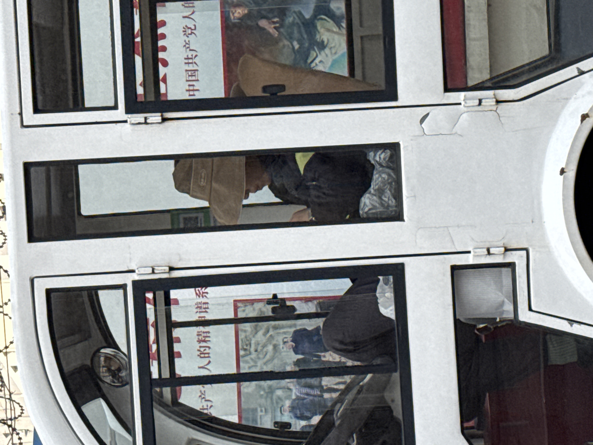A vertically oriented image depicts a double-decker bus, likely in Zhongtun, China. Through the bus windows, one can see passengers and posters. The top window has a poster in Chinese, possibly with a figure depicted. The middle window reveals a passenger wearing a hat and dark clothing. The bottom window contains another Chinese poster with multiple figures in it. The bus is white, and appears stationary. The presence of the posters indicates a public space, and their content implies a political or social message. The overall impression is of a candid snapshot of urban life in China.