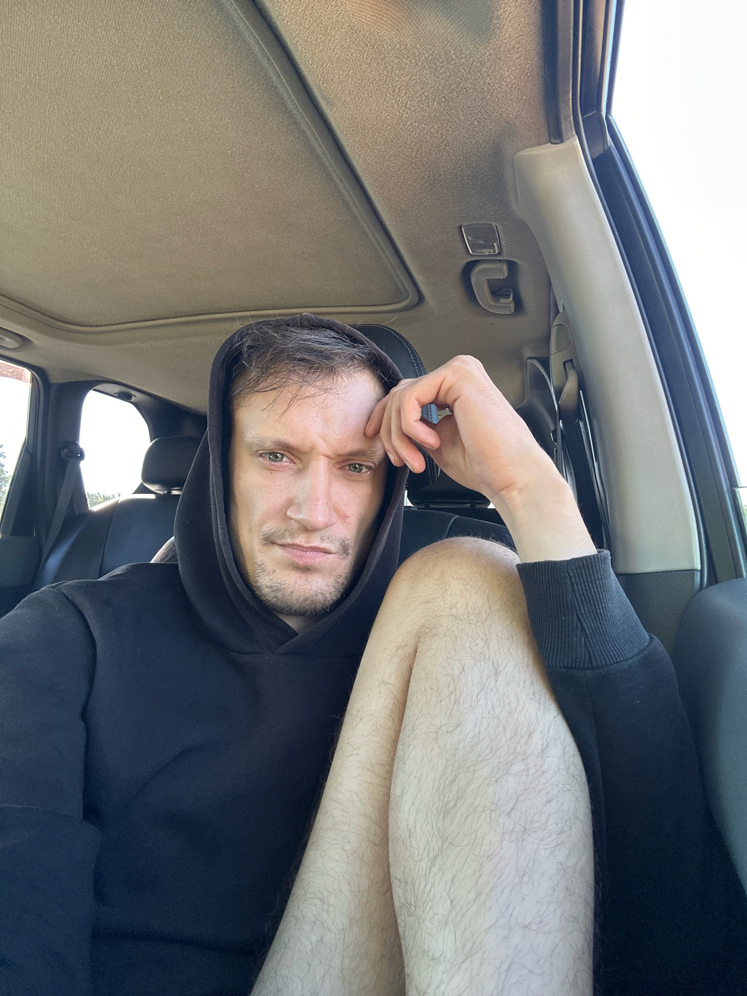 The image shows a man in a black hooded sweatshirt sitting in the driver's seat of a car. His left arm is resting on his leg, and his right hand is supporting his forehead. The car's interior is visible, including the sunroof and headliner. The man appears to be looking directly at the camera with a serious expression. The background shows a blurred view of trees and sky through the car window. The scene is set in an urban environment, likely Makiv, Ukraine, as indicated by the context.