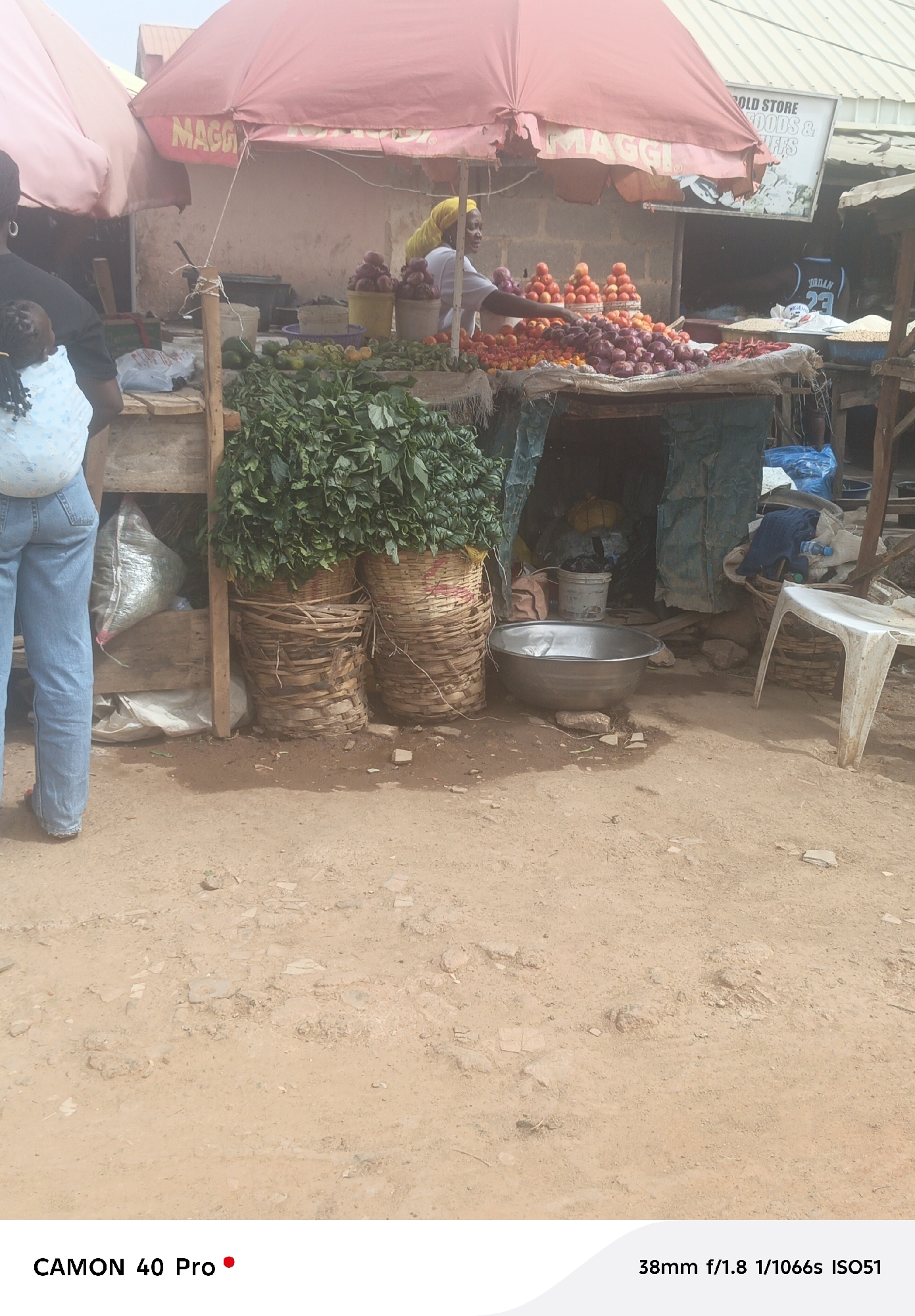 This photograph captures a vibrant open-air market stall in Jos, Nigeria, during what appears to be a bright daytime. The scene is dominated by a produce seller, a woman in a white top and yellow headscarf, standing behind a wooden stall laden with fresh vegetables. She appears to be interacting with customers, possibly the person seen from behind in the foreground, carrying a baby on their back in a patterned cloth, wearing blue jeans.
The market stall is shaded by two large, somewhat worn, red umbrellas, one bearing the "MAGGI" brand logo. On the stall, there are abundant piles of red onions, ripe tomatoes, and red peppers. Beneath the main display, large woven baskets overflow with green leafy vegetables. Other produce like limes or lemons are also visible. On the ground in front of the stall sits a large silver metal bowl, and a white plastic chair is positioned to the right. The ground is unpaved dirt, typical of outdoor markets.
In the background, behind the stall, stands a building with a "OLD STORE FOODS & STUFFS" sign. Another individual is visible in the background, wearing a black jersey with "JORDAN 23" printed on it. The overall impression is one of a busy, functional local market, offering fresh produce in a sunlit environment.