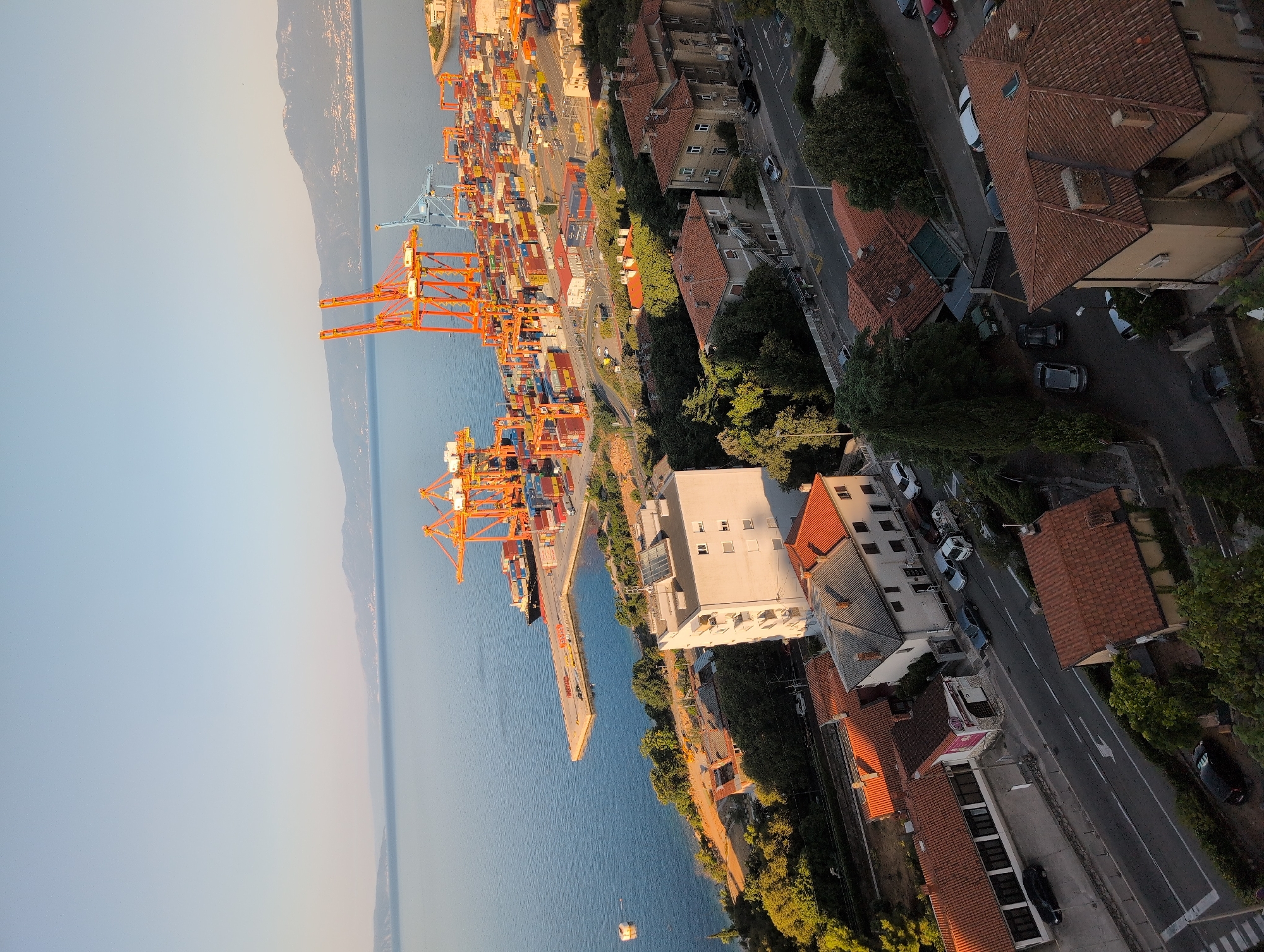 This is a high-angle, eye-level, medium shot of a port city. In the distance, a large cargo ship is docked at a pier filled with shipping containers and cranes. The sea is calm and reflects the clear blue sky. In the foreground, several residential buildings with red tiled roofs are nestled amongst lush green trees. A paved road winds through the neighborhood, with cars parked along the sides and driving on it. The overall scene depicts a bustling port integrated with a residential area. The location is the city of Grad Rijeka, Croatia, specifically the port area and surrounding residential hills overlooking the Adriatic Sea.