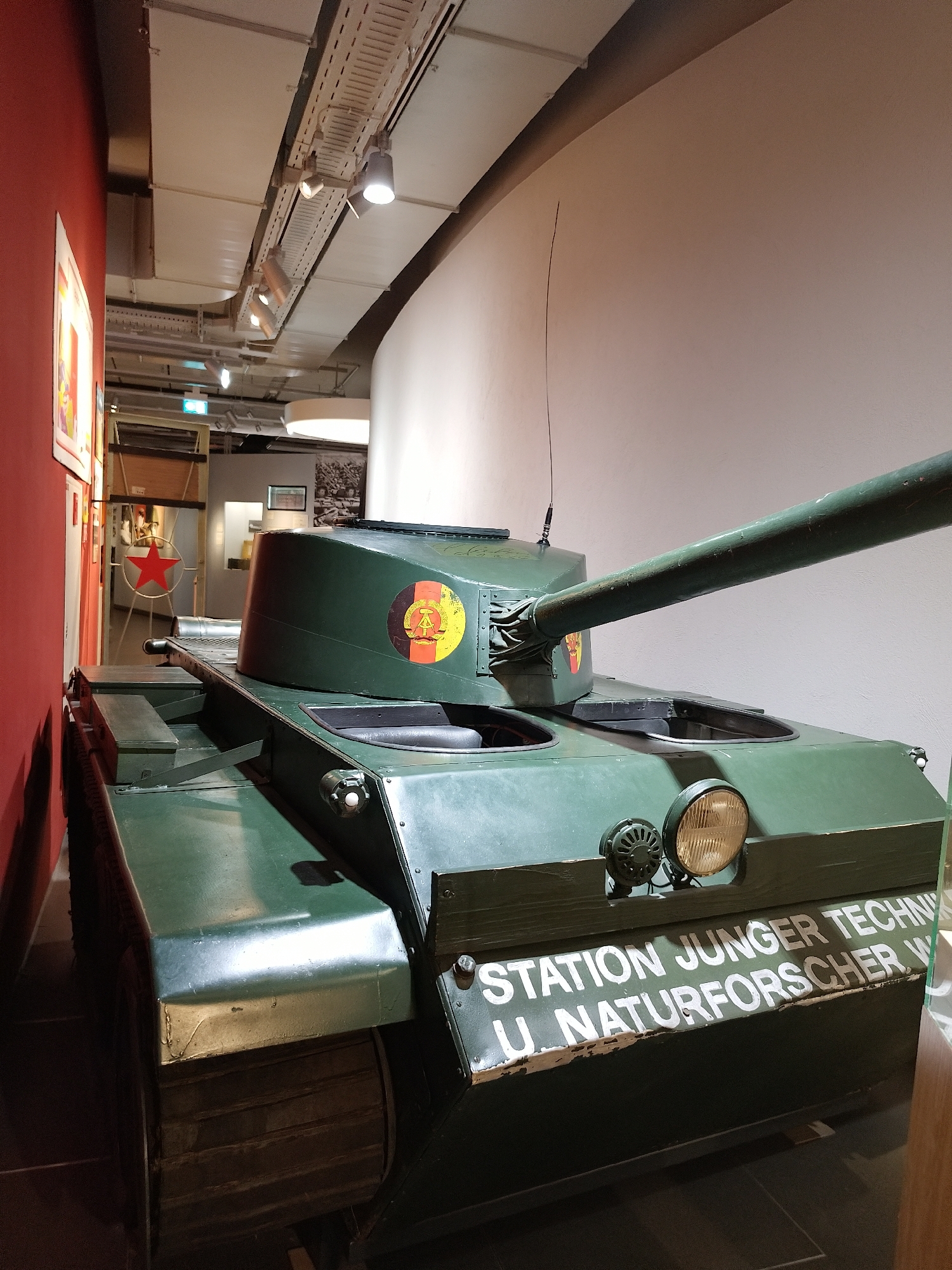 A green tank with an emblem of the German Democratic Republic is on display indoors. The tank is parked near a red wall with pictures, and a doorway with a red star is visible in the background. The tank has lettering that reads "STATION JUNGER TECHN. U. NATURFORSCHER." The location appears to be a museum, based on the artifacts and display setup. The city is Unknown, Unknown.