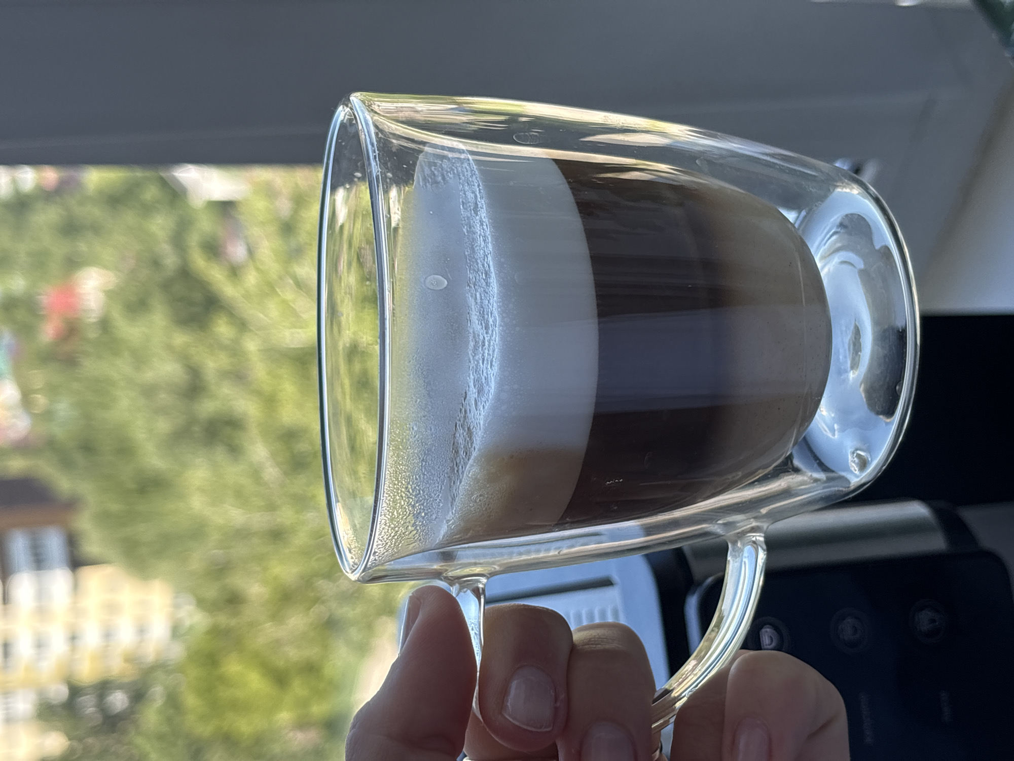 A hand, appearing to be a person's, holds a clear, double-walled glass mug filled with a layered beverage, possibly coffee with milk or a similar drink, featuring distinct bands of dark liquid and frothy white foam. The mug has a curved handle. Behind the mug, out of focus, is a blurry green natural environment with trees, and beyond that, a blurry building with yellow and white elements, suggesting an urban setting. To the right of the mug, a gray, vertical element is visible, possibly a window frame or a wall. The scene is illuminated by natural light, casting reflections on the glass mug. The perspective is close-up, focusing on the mug and the hand. The background imagery suggests an outdoor view, possibly from an apartment or office building. The location in the city of Perm, Russia, cannot be definitively determined from the visual information alone, as the background elements are too indistinct.