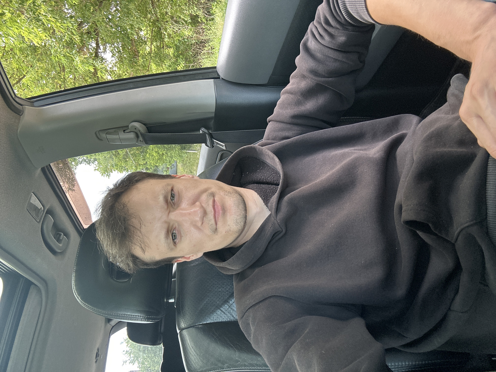 A man, appearing to be in his late 20s or early 30s, is seated in the driver's seat of a car, taking a selfie. He has light skin, short brown hair, and a light stubble. He is wearing a dark grey hooded sweatshirt. The car's interior is visible, including the black leather driver's seat, a sunroof, and the car's door frame and window. Outside the car, lush green trees and foliage can be seen, suggesting a rural or semi-rural setting. The image is taken from a slightly high angle, looking down at the man. Based on the exterior vegetation and the visible style of the car, this scene could plausibly be located in or near Makiv, Ukraine, a region characterized by its natural beauty and agricultural landscapes.