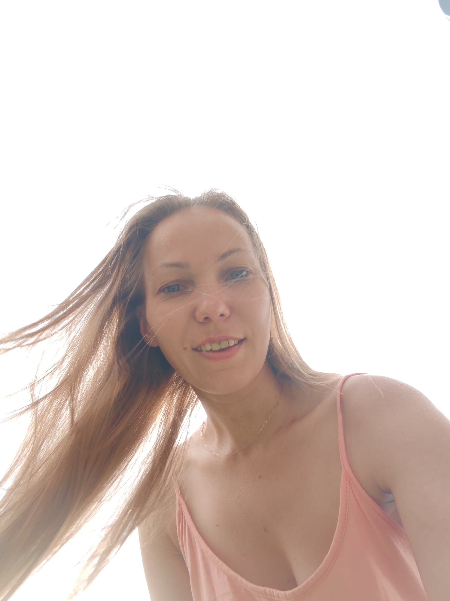 A low-angle selfie shows a woman from the chest up against a bright, overcast sky. Her long, light brown hair is blowing to the left, obscuring the left side of her face. Her head is tilted slightly to the right, and she is looking towards the camera with a subtle smile. She is wearing a pink tank top. The overall lighting suggests an outdoor setting, likely during the day, possibly in a park or open area. The sky is uniformly bright, indicating an overcast or very hazy condition. The image does not contain any identifiable landmarks or context that would allow for a specific location within Чухлэм, Russia to be determined.
