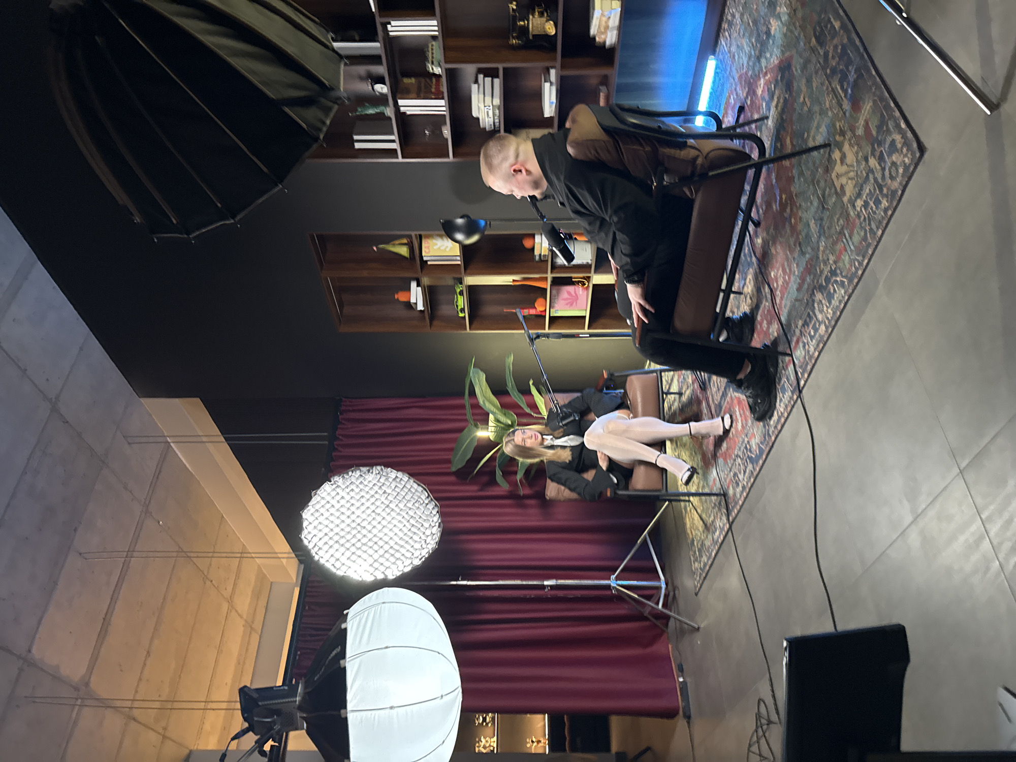 The image depicts an interior scene, likely a studio, in Warsaw, Poland. A man with a shaved head, dressed in black, sits in a leather chair, leaning towards a microphone. A woman lies on a leather couch, facing the camera, wearing a dark blazer, a white shirt, and white tights. Both are likely involved in some kind of recording or performance.
Behind them are dark red curtains and a large potted plant. There are two bookcases filled with various objects and books. Studio lighting equipment, including a large umbrella reflector and a softbox, are visible in the foreground. An oriental-style rug covers part of the floor. A monitor or TV screen is placed in the lower right of the image. The floor is tiled.