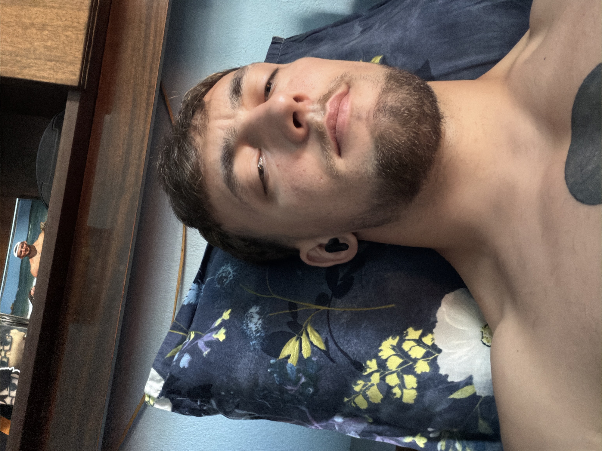 The image shows a close-up, selfie-style shot of a man lying on a pillow. The man has short brown hair, a beard, and is wearing black wireless earbuds. He is shirtless, revealing his chest and collarbone. The pillow has a dark blue floral pattern with yellow and white flowers. In the background, a wooden shelf is visible, with framed photos on it. One photo shows a man in a white baseball cap on a beach. The wall behind the man appears to be light blue. The scene is indoors, likely in a bedroom.
