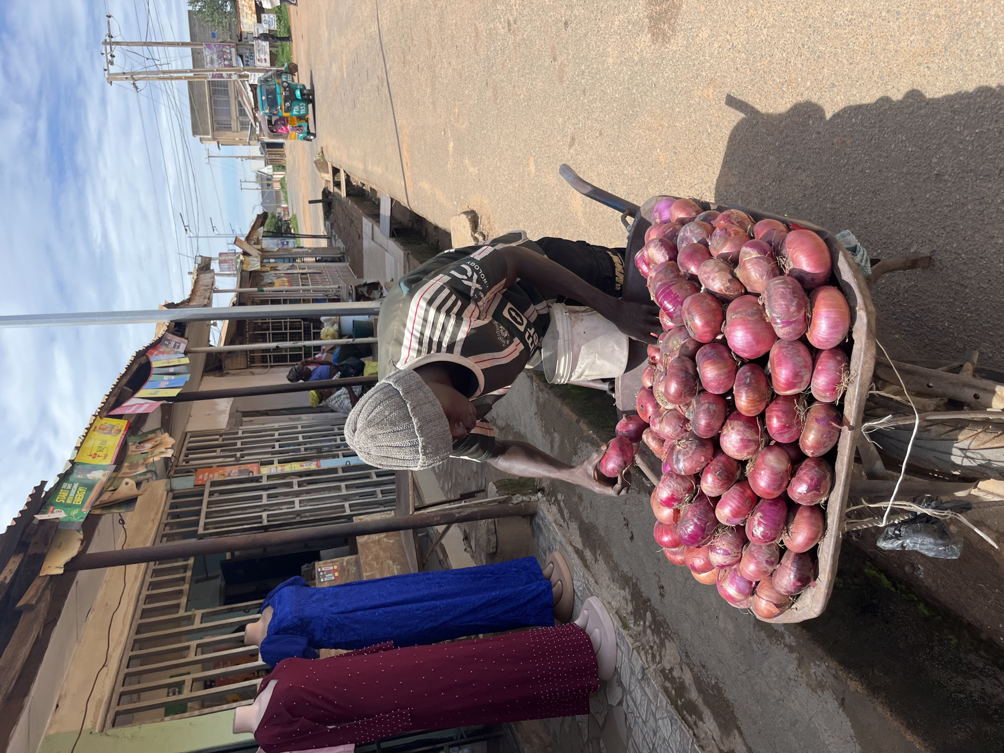 An individual wearing a grey knit beanie, a dark t-shirt with striped sleeves, and dark trousers is observed bending over a metal wheelbarrow filled with numerous red onions. The person is holding a single red onion in their left hand, seemingly engaged in sorting or selecting produce. A white plastic bucket is positioned near their right hand. The wheelbarrow rests on the edge of a paved road.
To the left, a row of single-story commercial buildings, featuring corrugated metal roofs and security-barred windows and doors, line a concrete sidewalk. Two mannequins displaying full-length dresses, one maroon with white dots and the other royal blue, are visible outside one of the storefronts. Various banners, some weathered, are affixed to the building overhangs, with one clearly stating "START BUILDING ENERGY!". A narrow, unpaved drainage channel runs along the edge of the sidewalk before meeting the asphalt road.
The asphalt road extends into the background under a partly cloudy sky. Utility poles carrying power lines are visible along the roadside. Further down the street, a green and yellow motor tricycle, common in Nigeria and known as a 'keke napep', with "PTA 105-" visible on its side, is either stopped or in motion. Additional buildings are present in the distance on both sides of the road, and faint figures of other people can be seen. The scene is illuminated by bright sunlight, casting distinct shadows on the ground. This depiction is consistent with a bustling market street environment in an urban area, specifically in Jos, Nigeria.