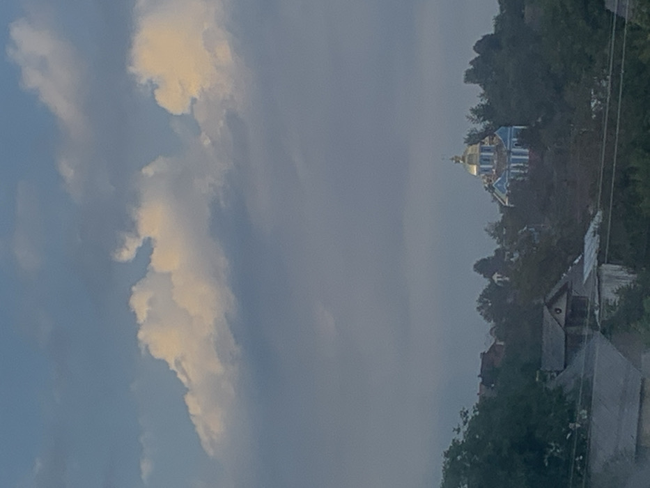 The image displays a daytime view of the Church of the Dormition of the Blessed Virgin Mary in Deliatyn, Ukraine. The church is positioned prominently on a tree-covered hill in the midground, featuring a central golden dome and light blue walls with white architectural details. Dense green foliage surrounds the church, with other residential buildings partially visible among the trees on the hillside. In the foreground, the upper sections and dark roofs of additional structures are discernible, partially obscured by more dark green foliage. Horizontal lines, consistent with utility wires, traverse the lower portion of the frame. The sky occupies the upper half of the image, presenting a mix of light blue and various cloud formations, including bright, sunlit cumulus clouds in the upper sections and more muted, greyish clouds filling the expanse below them. No individuals or active interactions are visible within the scene.