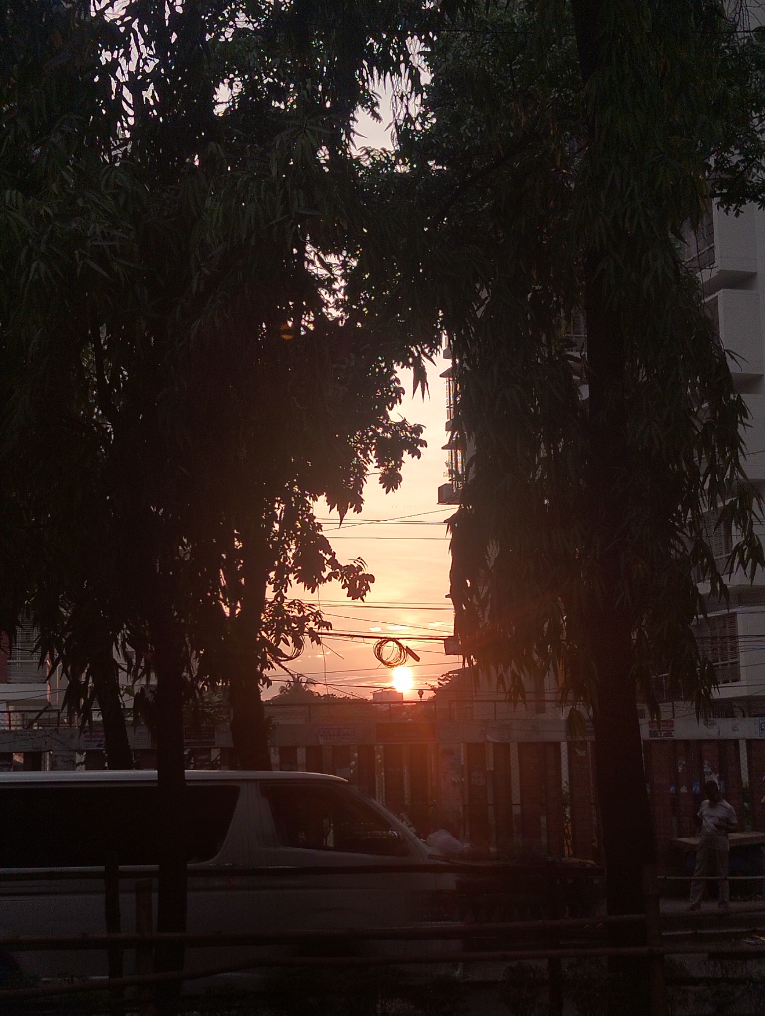The image shows a street scene in Dhaka, Bangladesh at sunset. Silhouetted trees frame a view of the setting sun, which is partially obscured by buildings. A white van is parked in the foreground, and a person stands on the sidewalk near a building with vertical bars. Utility wires are visible against the sunset sky. The overall scene is dark with the exception of the bright setting sun.