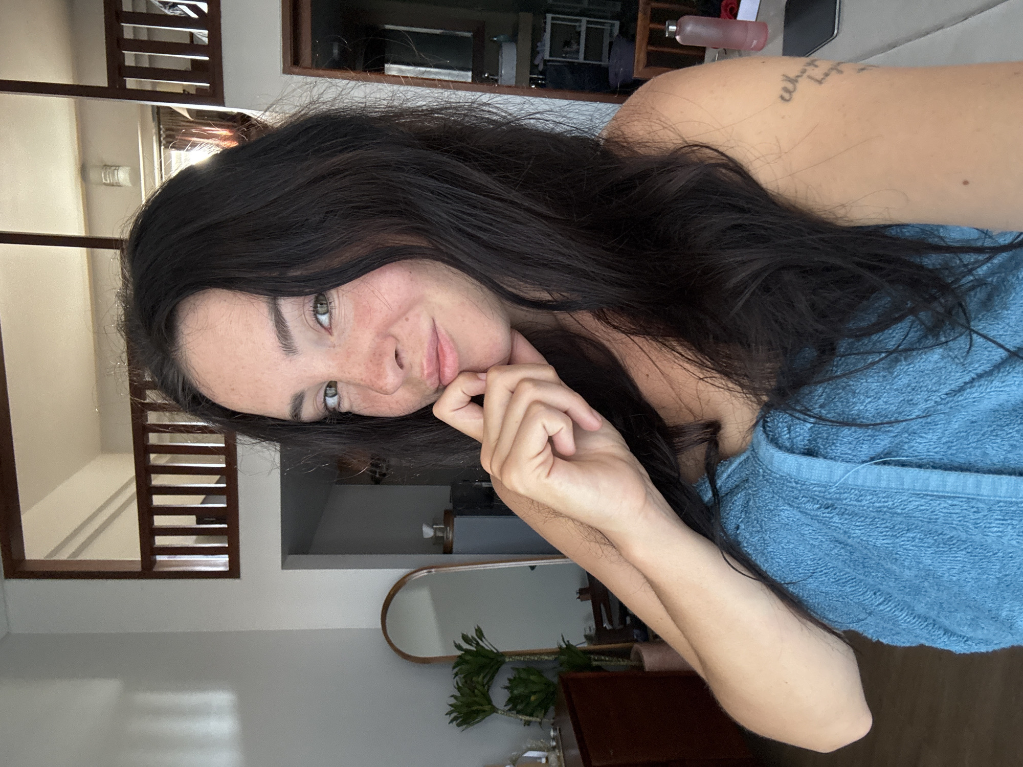A woman with dark hair and freckles is taking a selfie in what appears to be a multi-story apartment or home. She is wrapped in a blue towel and has her hand on her chin. Behind her, a wooden railing suggests an upper level with a view of the ceiling. To her left, a tall, oval mirror reflects a dresser and a potted plant. The room is lit by natural light, casting shadows on the wall. The overall impression is a casual, personal moment captured in a domestic setting.