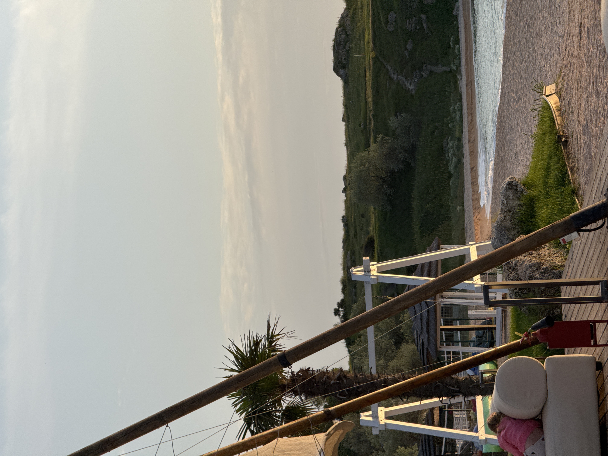 The image shows an outdoor scene in Мысовское сельское поселение, Ukraine. A beach is visible with waves gently rolling onto the shore. A grassy hill rises behind the beach. In the foreground, there's a wooden structure with white supports, likely part of a beachside establishment. A palm tree is positioned near the structure. A partially visible white sofa with a person in pink partially hidden by it rests on the wooden deck, along with a red object. The sky is overcast.