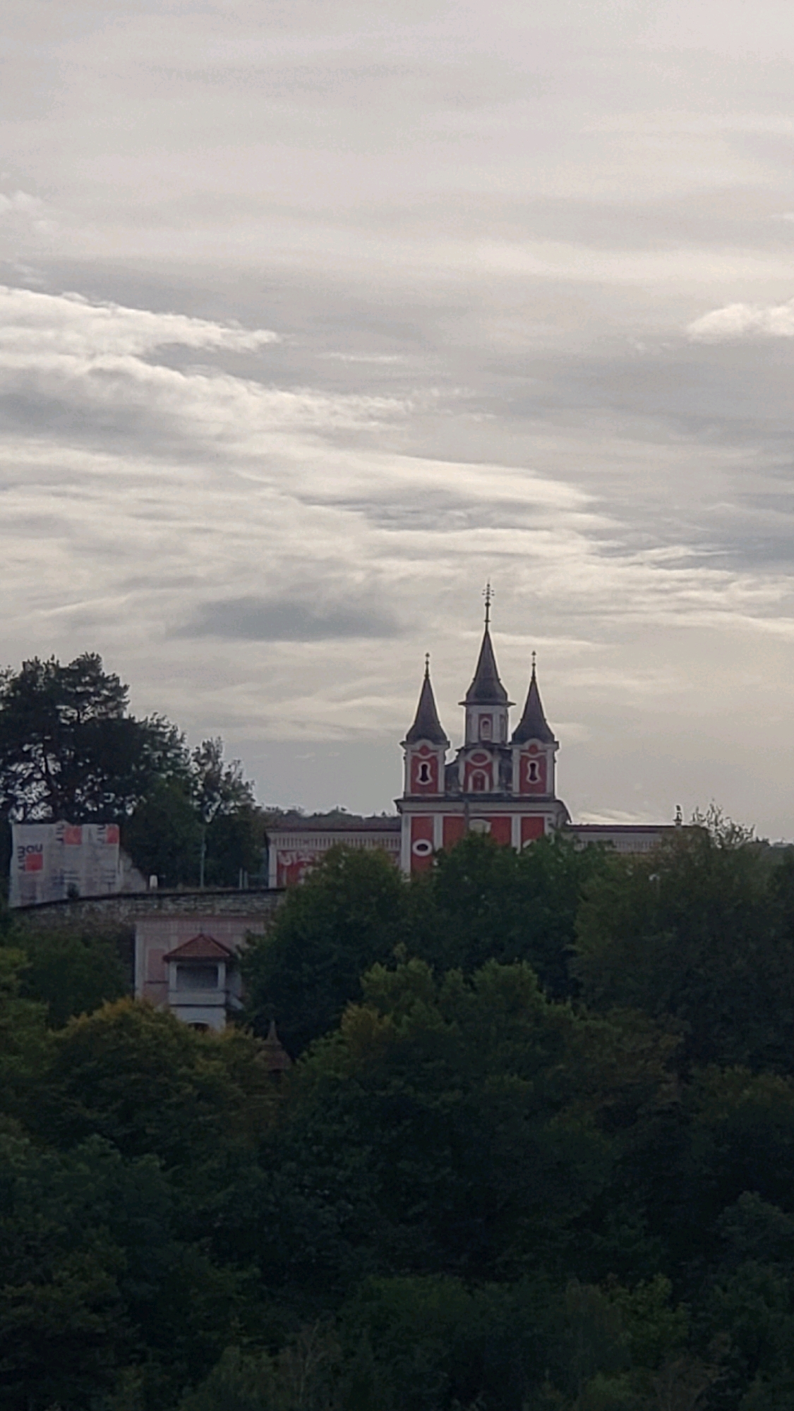 The image shows a church with three spires, painted red and white, set against a cloudy sky. The church is surrounded by lush green trees. In the foreground, there are more trees, and in the background, there is a wall under construction with some building materials. The church is likely the Orthodox Church of St. John the Baptist in Prešov, Slovakia, known for its distinctive architecture and prominent position within the city. The overall scene depicts a serene, somewhat overcast day, with the church standing out as a significant landmark.