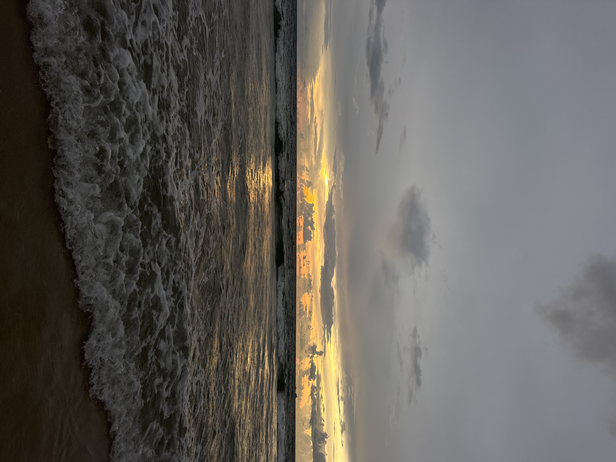 A twilight view of the Andaman Sea from the shore in Choeng Thale, Thailand, shows a dramatic sky with dark, heavy clouds interspersed with patches of yellow and orange light near the horizon. The sea is dark and choppy, with white foam from breaking waves reaching the sandy shore in the foreground. The reflection of the sunset light can be seen on the water's surface.