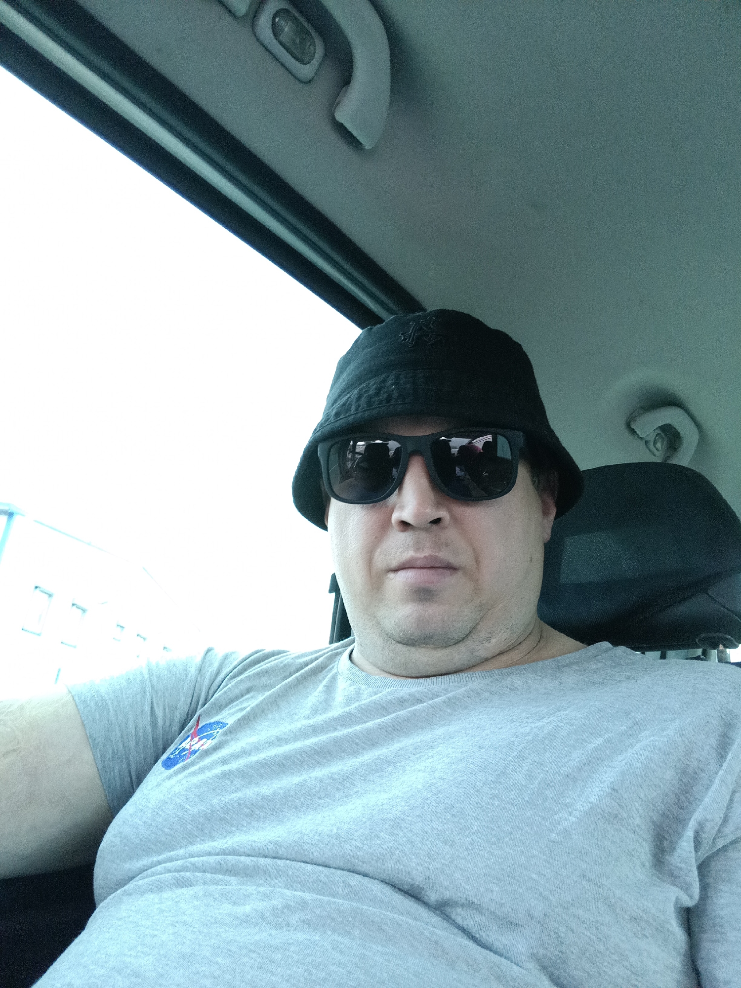 The image presents an adult male seated within the passenger compartment of a vehicle in the city of Dalovy, Czechia. The individual, of stocky build and fair complexion, occupies the foreground, looking directly towards the camera. He wears a black bucket hat with an embroidered logo and black-framed sunglasses, which obscure his eyes. His mouth is closed, maintaining a neutral expression, and visible folds are present on his neck. He is dressed in a light grey short-sleeved t-shirt featuring a blue, red, and white NASA logo on the left chest. The vehicle's interior is visible, including a light grey/teal-grey headliner, an overhead light fixture in the upper right, and a black headrest behind the subject's right side. Through the vehicle's left window, an extremely bright, overexposed outdoor scene is visible, with faint, distant architectural outlines suggesting buildings under strong daylight conditions.
