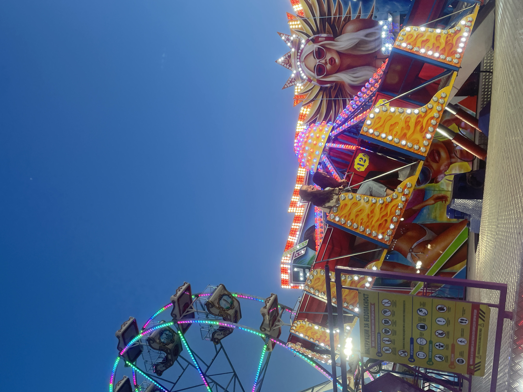The image shows a Ferris wheel and a brightly lit carnival ride with a woman's image and flames. The ride appears to be in motion. Safety guidelines are visible on a sign in the foreground. The sky is clear and blue. The location is Unknown, Unknown.