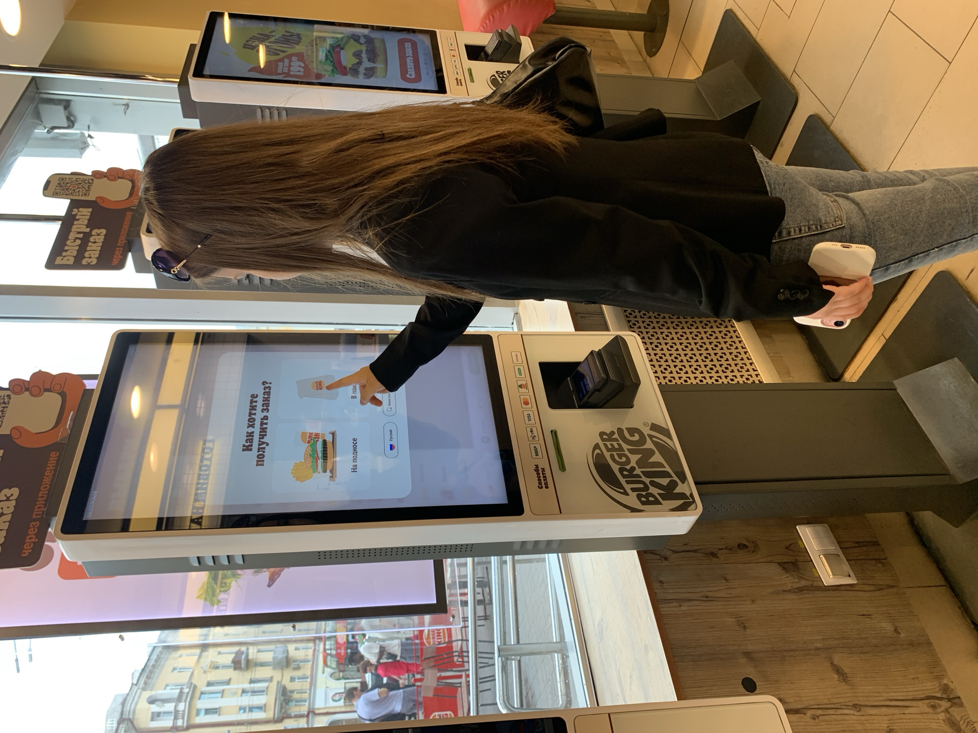 A young woman with long brown hair, wearing a black blazer and blue jeans, stands at a Burger King self-service kiosk in Kazan, Russia. She is holding a white smartphone in her right hand and appears to be interacting with the touchscreen. The kiosk's screen displays options in Russian, with the question "How do you want to receive your order?". Two options are visible: "For pickup" with an image of a burger and fries, and "In a bag" with a Burger King bag. Another kiosk is visible to the right, and through a large window to the left, a street scene with buildings, people, and a red double-decker bus can be seen. The overall environment suggests an indoor fast-food restaurant.