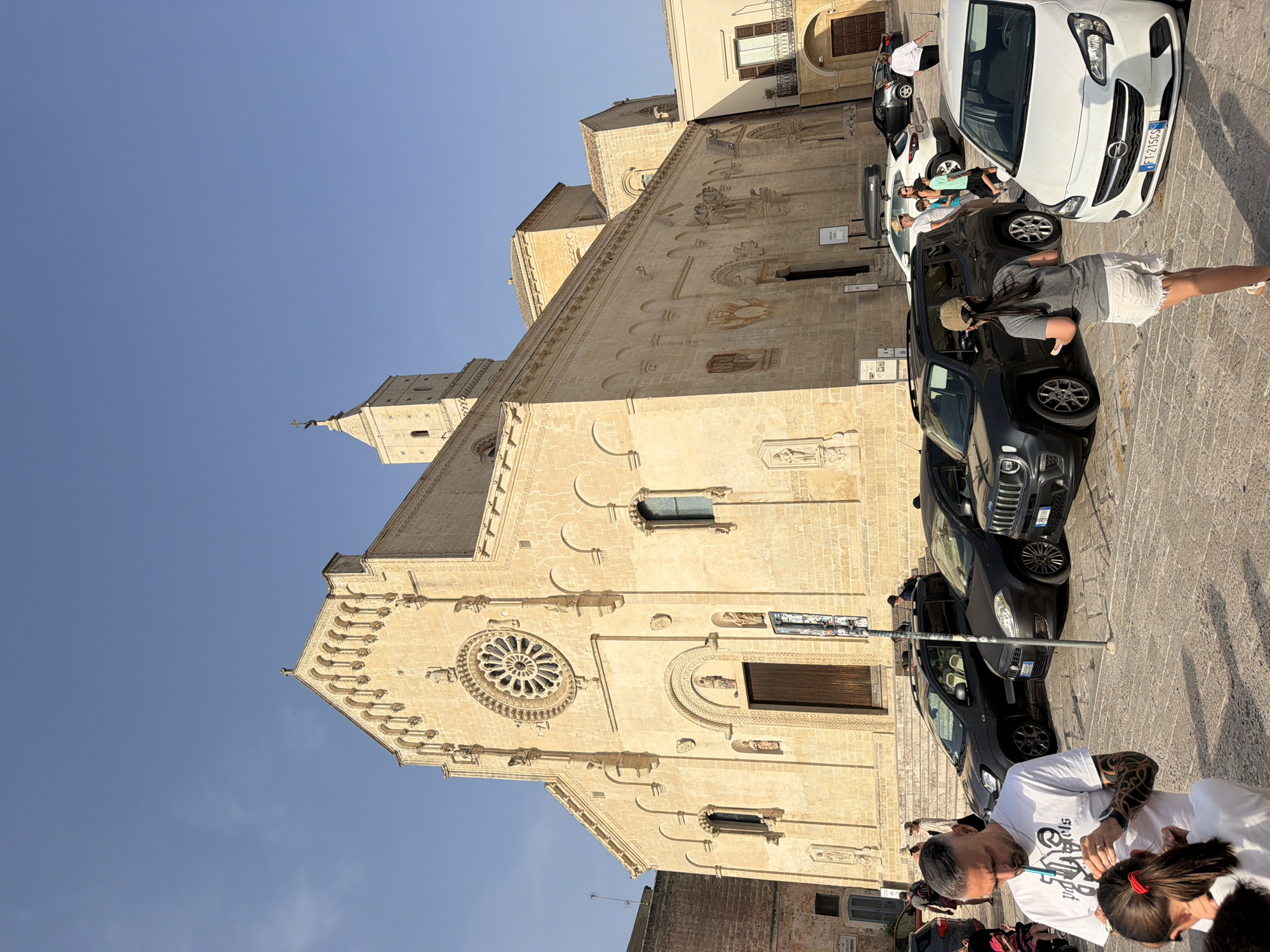 The image captures a view of the Matera Cathedral, also known as the Cattedrale di Maria Santissima della Bruna, located in Matera, Italy. The cathedral is a large, ancient stone building with a prominent rose window on its facade and a bell tower. The sky above is clear and blue. In the foreground and surrounding the cathedral, there is a cobblestone plaza with several cars parked. People are visible in the plaza, some walking, some interacting, and some attending to their vehicles. Among them is a man with tattoos on his arms, wearing a white t-shirt, and a young girl with her hair in a ponytail. Another woman, wearing a cap and shorts, is seen from behind, walking towards the foreground. The overall scene depicts a sunny day in a public space adjacent to a historic religious structure, with everyday activities taking place around it.