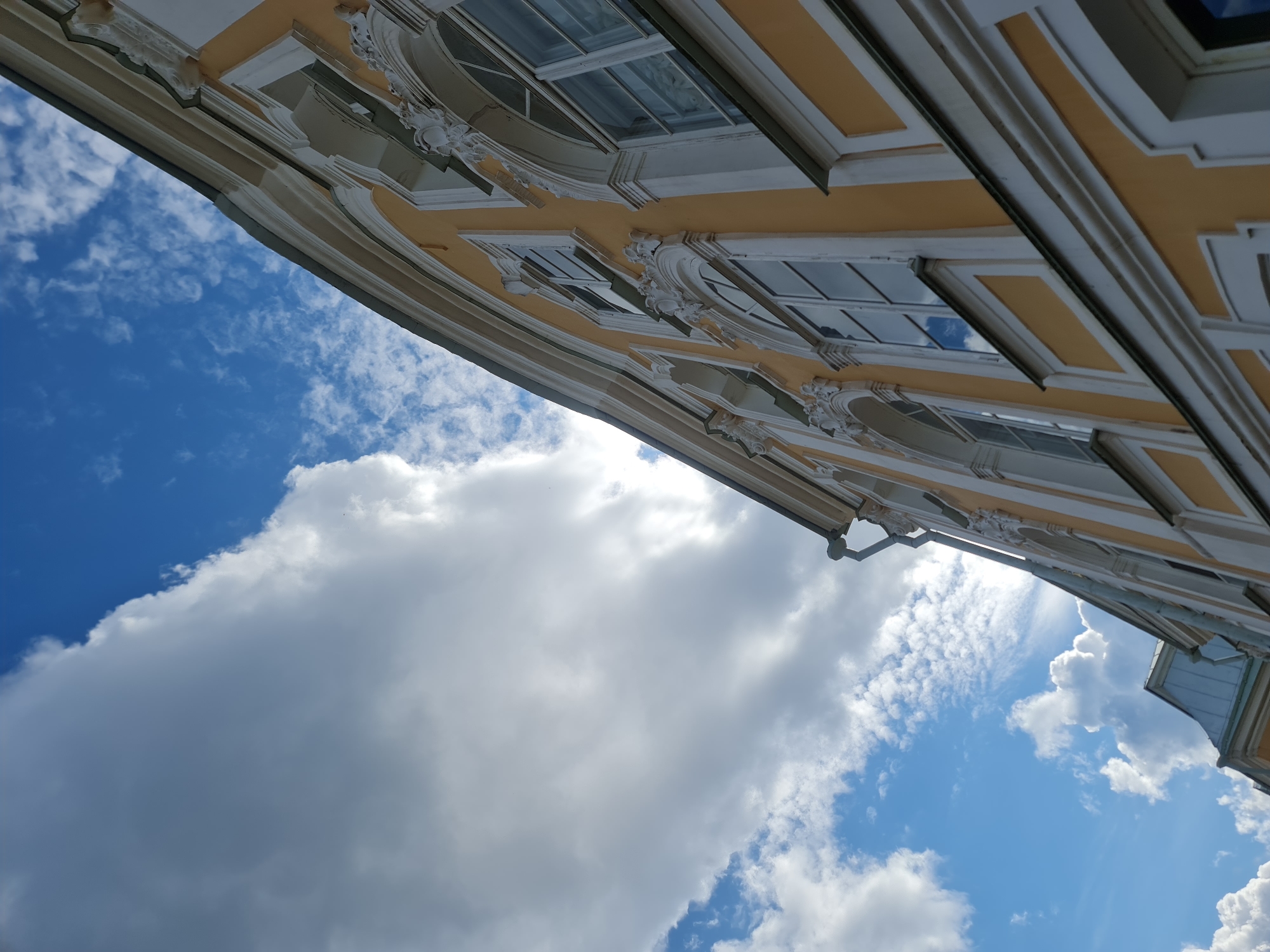 The image captures a low-angle view of the facade of a yellow building with white ornate trim, located in Peterhof, Russia. The building features several arched and rectangular windows, some reflecting the sky. Above the building, a dramatic sky is visible, filled with large, billowing white clouds against a bright blue backdrop. The lighting suggests it is daytime, with sunlight filtering through the clouds and illuminating parts of the building and sky. The architectural style appears to be classical or baroque, characteristic of historic buildings in Peterhof. No people or specific actions are depicted, and the scene is static.
