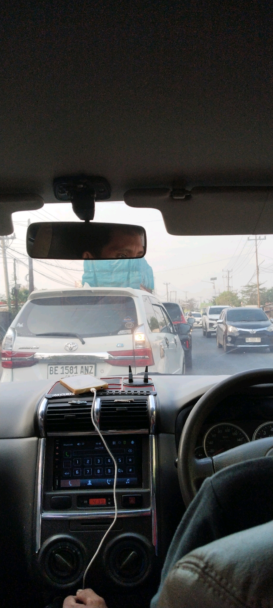 The image shows the interior of a car, looking out through the windshield. The car is in heavy traffic on a paved road, with other vehicles visible ahead. The dashboard of the car is prominent, featuring a car stereo with a touchscreen display, air vents, and a phone charging on a mount. A license plate on the car in front reads "BE 1581 ANZ". The scene appears to be in a city, possibly Terbanggi Besar, Indonesia, given the context provided. The sky is overcast, suggesting it might be daytime. The rear-view mirror reflects the face of the driver, who is looking forward.