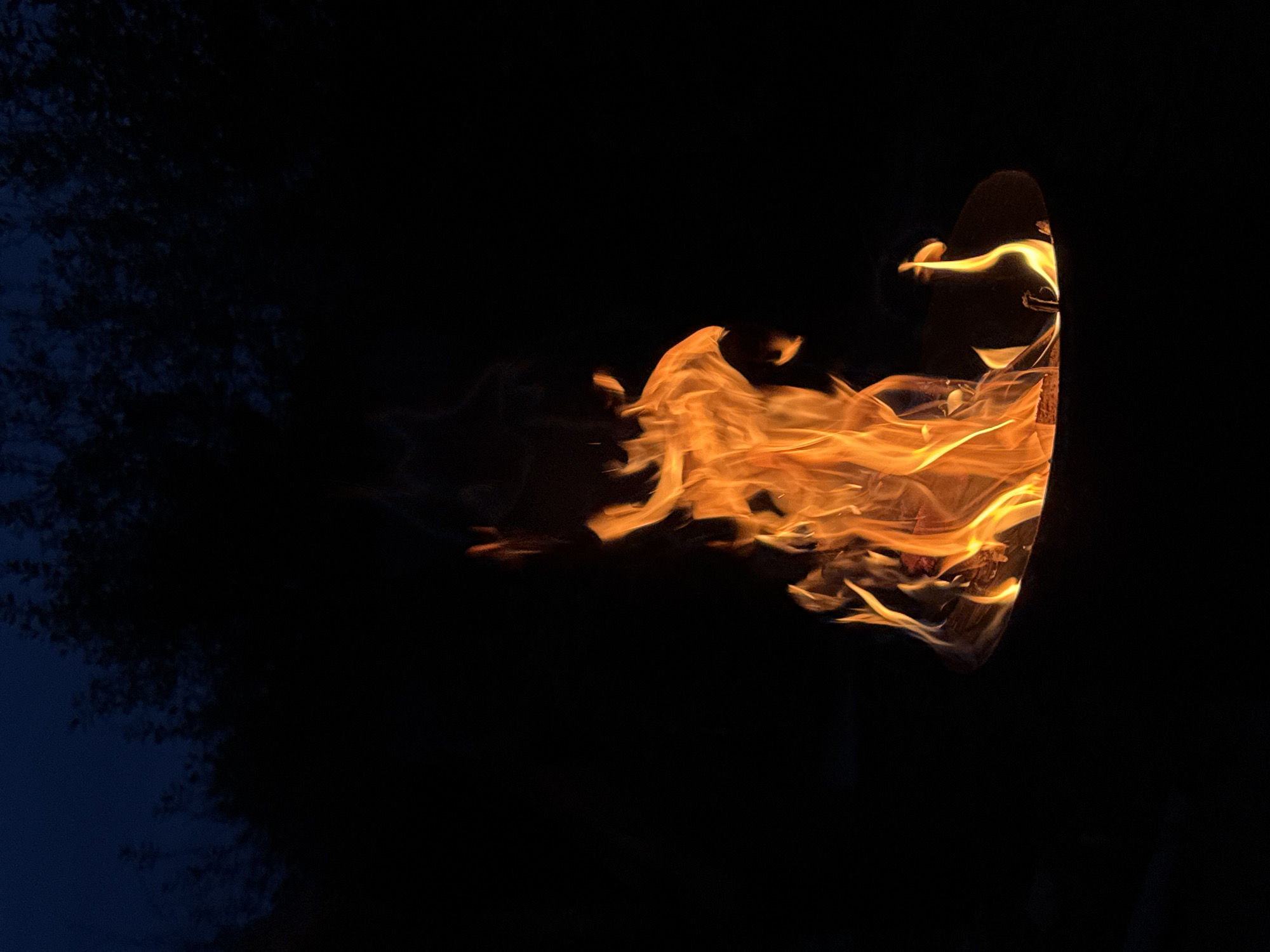 A campfire burns brightly at night, with flames reaching upwards against a dark background. The fire is contained within a metal fire pit. In the background, the silhouettes of trees are visible against a deep blue, twilight sky. The scene is dark, with the fire providing the primary source of light. The location in Chaikovsky, Russia, is not discernible from the image itself.