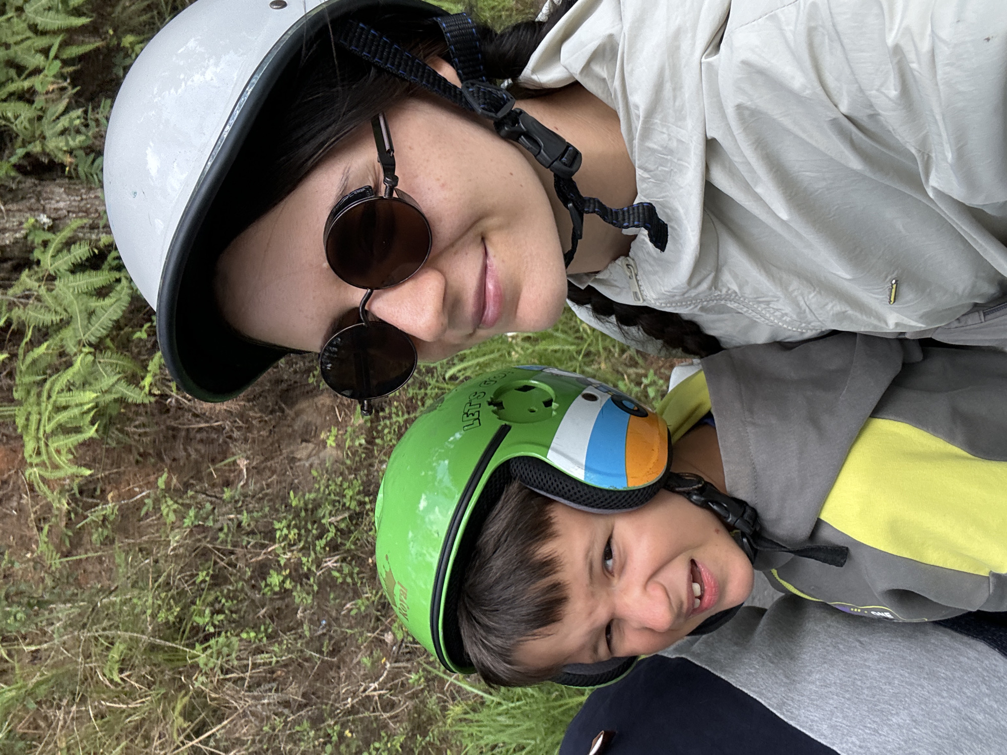 Two individuals, an adult woman and a child, are captured in a close-up, upward-angled shot against a natural outdoor backdrop, likely taken in Da Lat, Vietnam. The original image is oriented sideways, with subjects appearing to lie horizontally. Both subjects are wearing helmets, suggesting an activity like cycling or motorbiking.
The woman, positioned higher in the frame, wears a white open-face helmet with a black trim and dark, round-framed sunglasses. She has dark hair, partially visible as a braid, and a subtle smile. She is dressed in a light-colored, possibly beige or light grey, lightweight jacket. A small yellow rectangular detail, resembling an "i" or "j" logo, is visible on the lower right side of her jacket.
Below her is a child, wearing a bright green helmet featuring colorful cartoon-like elements on the front, including blue, orange, and white stripes, possibly depicting eyes. The helmet also has text that reads "LET'S GO" on the top and "ROYAL" with a small crown symbol on the side. The child has short, dark hair and an expressive, somewhat playful or grimacing facial expression with teeth visible. They are wearing a multi-toned hoodie in shades of grey, yellow, and navy blue, with partially visible text, possibly "ONE," on the chest area.
The background consists of natural ground cover, including green ferns, dry grass, small green plants, and earth, indicating an outdoor, possibly wooded or park-like setting. The lighting suggests it is daytime, with soft, indirect light.
