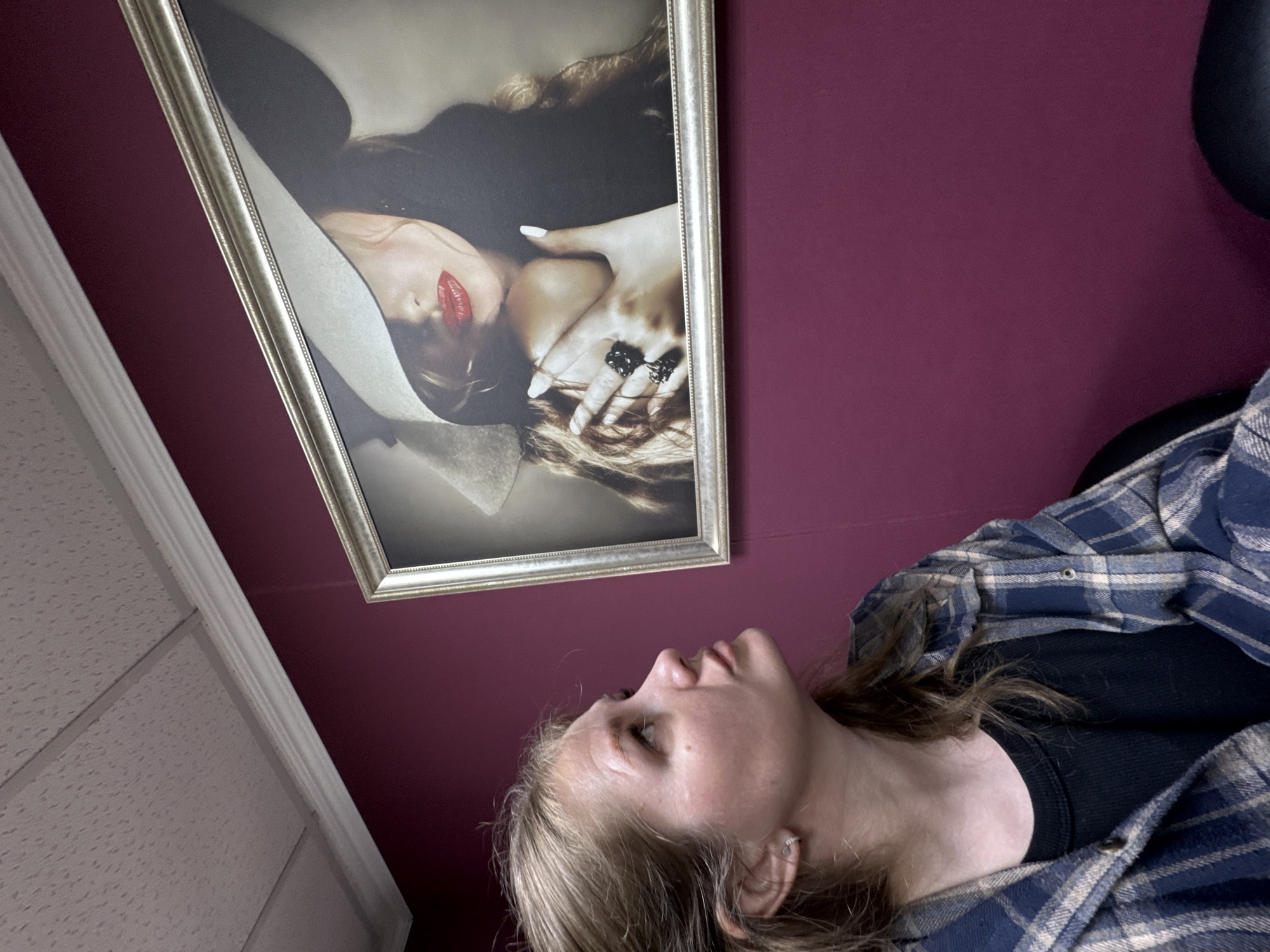 A person wearing a blue and tan plaid shirt and black shirt is looking up at a framed picture hanging on a maroon wall. The picture is of a woman with red lipstick, a white hat, and a hand with rings on it near her face. A textured ceiling is visible in the upper left corner of the image. The location is unknown, and there is no indication that the image was taken in Perm, Russia.