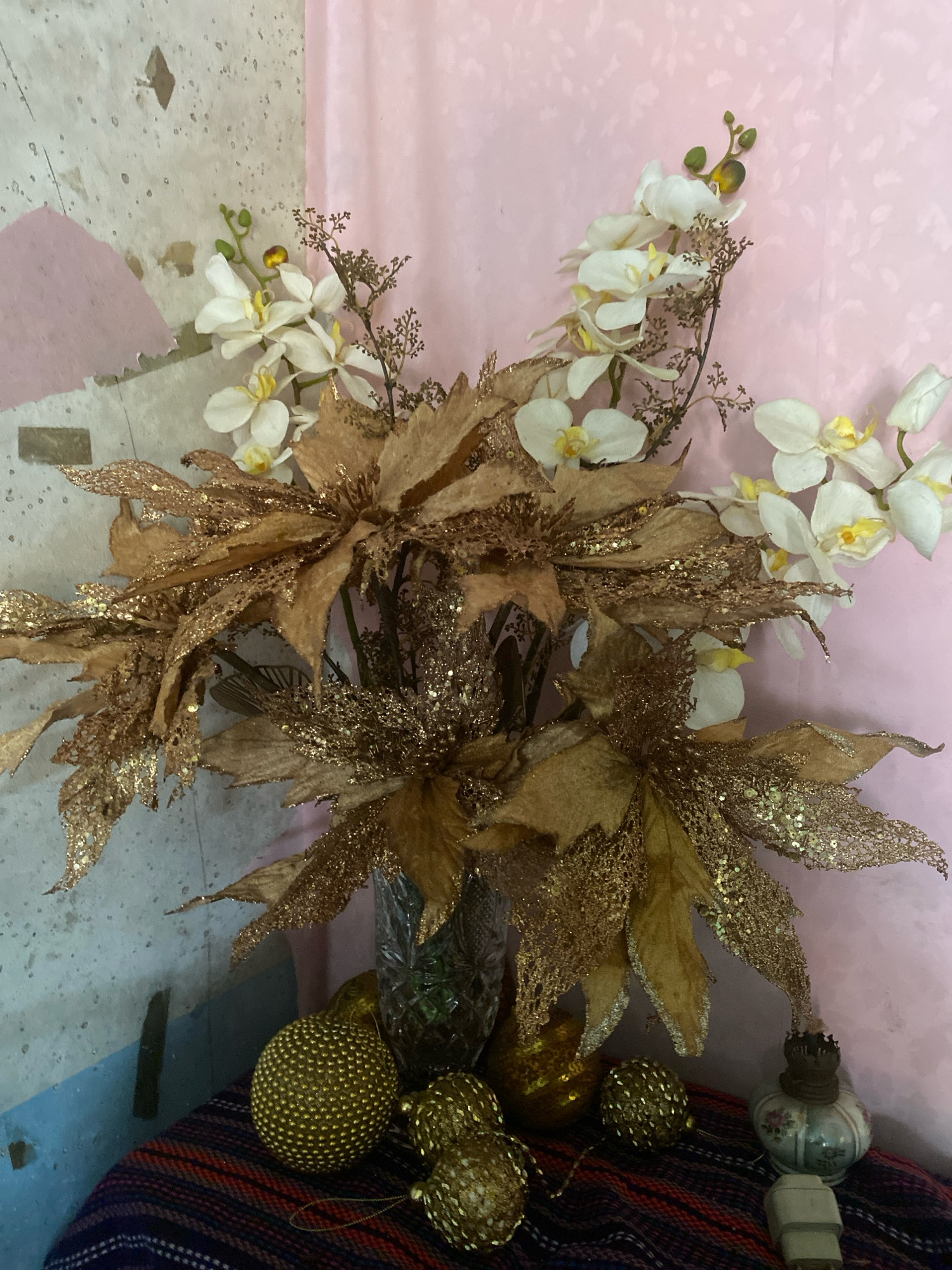 A floral arrangement featuring artificial white orchids and gold glitter-covered poinsettia-like flowers in a clear glass vase is displayed on a table covered with a striped cloth. Several gold decorative spheres and a small antique-style oil lamp with a floral pattern are placed around the vase. The background consists of a textured wall with pink and blue accents on the left and a pink patterned fabric on the right. The scene appears to be an indoor setting. No people are present.