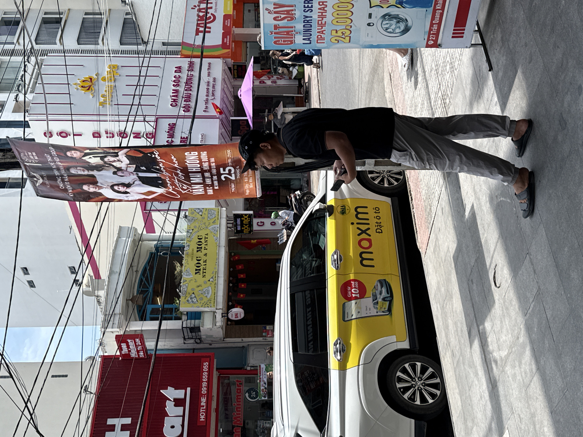 A pedestrian views a cell phone next to a white and yellow Maxim taxi in Nha Trang Ward, Vietnam. Street-level store signage and advertising posters dominate the background. The street scene includes power lines and adjacent buildings.