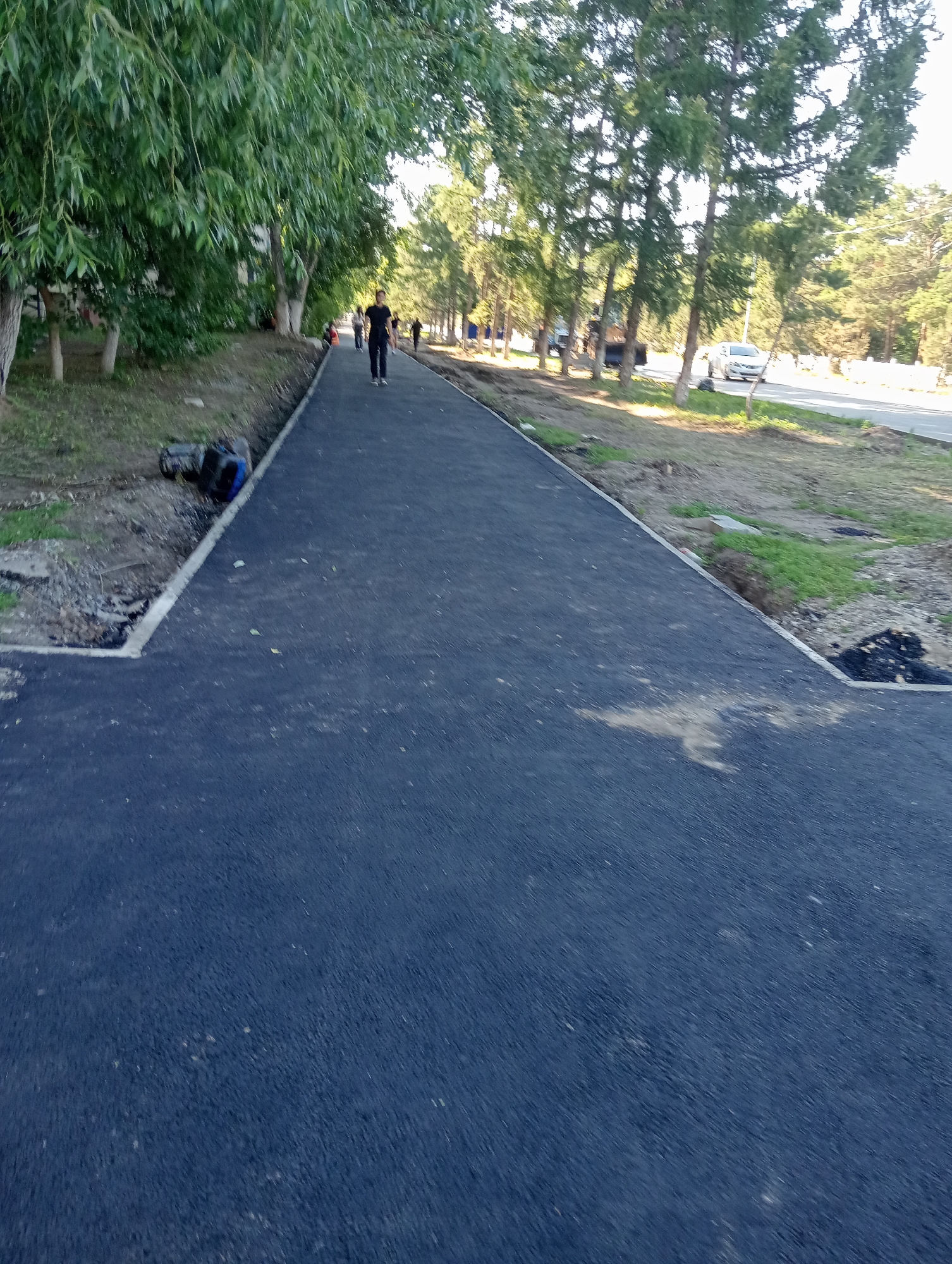 A new asphalt path has been laid along a tree-lined street in Kostanay, Kazakhstan. The path is dark and appears freshly paved, with a white concrete curb on the left side. To the left of the path, there is a grassy area with some trees. Further left, a person is walking on the path, wearing dark clothing. In the distance, several other people are also visible walking on the path. Across the street, there are more trees and a white car is driving by. The environment suggests a park or a residential area where infrastructure is being improved.