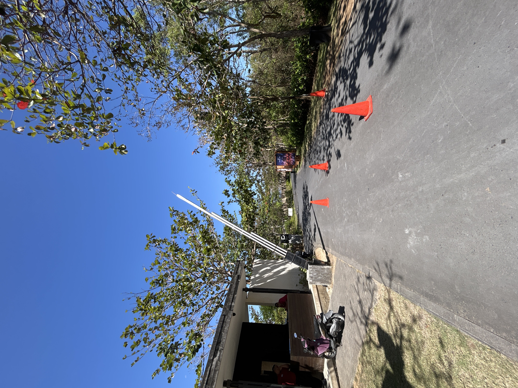 The image depicts a clear, sunny day at what appears to be an entrance to a property or complex in Kuta Selatan, Indonesia. A black and white striped boom gate is raised, suggesting entry is permitted or that it is currently open. Orange traffic cones are placed along the asphalt road, indicating a potential traffic management or temporary closure setup.
On the left side of the image, a small building with dark colored walls and a lighter colored roof overhang is visible. A purple scooter is parked on a paved area in front of this building. Two individuals wearing red shirts and dark pants are standing behind a counter area within the building, possibly security or staff members. Trees with green foliage line the sides of the road and surround the area, casting shadows on the ground. In the distance, more trees and vegetation are visible, suggesting a semi-rural or resort-like setting. The sky is a vibrant, cloudless blue. The overall scene conveys a sense of an outdoor access point on a bright day.