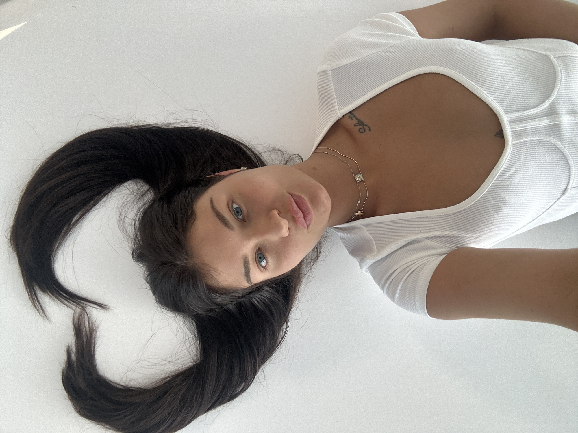 A young woman lies on a plain, light-colored surface, looking directly into the camera with her head tilted. Her long, dark hair is spread out around her head, forming a flowing shape against the white background. She has striking blue eyes and a neutral expression.
She is wearing a white, ribbed top with a wide scoop neckline. Around her neck, she wears a delicate gold chain necklace featuring a small square pendant and another smaller charm. Small stud earrings are visible.
Visible on her chest/shoulder area are two tattoos: one appears to be text, partially readable as "Sebastien," and another is a small, dark symbol near her collarbone. The background is uniformly white, suggesting an indoor setting with minimalist aesthetics. The lighting is soft and even. The scene takes place in Khimki, Russia.