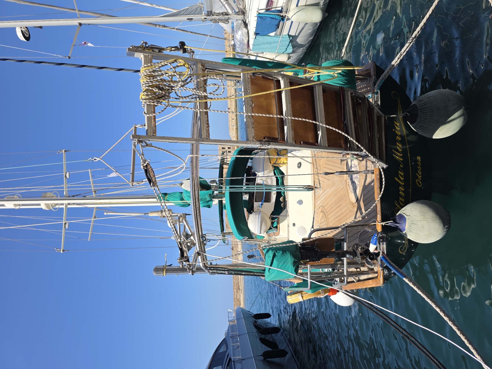 The image shows the stern of a sailboat named "Santa Maria Odessa" docked in the harbor of Chania, Greece. The sailboat is dark green with gold lettering and has a wooden deck. It is equipped with multiple ropes, green covers, and several buoys. To the left, a sleek, modern white yacht is docked. To the right, another sailboat is visible, with blue and turquoise towels hanging from its side. The water is clear and blue, reflecting the clear blue sky. In the background, a breakwater made of stone separates the harbor from the sea, and a few buildings are visible on the coastline. The scene suggests a calm, sunny day in a maritime setting.