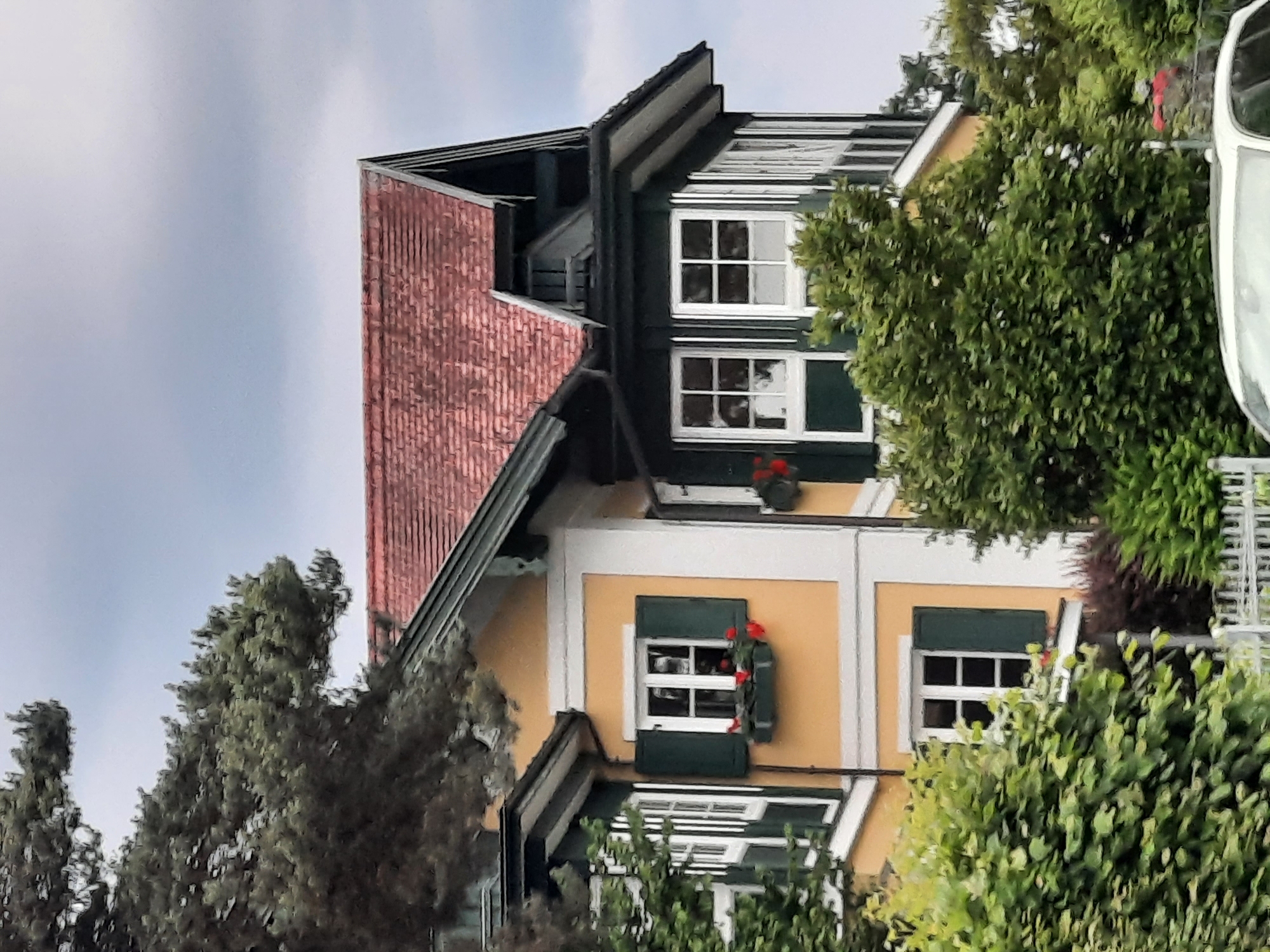 The image shows a section of a building, likely a house, with a yellow facade and a red tiled roof. It is partially obscured by lush green foliage and a large tree with dark green needles in the foreground. The building features multiple windows with dark green shutters and white trim. Some windows have flower boxes with red flowers. The style of the architecture appears to be traditional European. The sky is overcast with a muted gray tone. The image is captured from a medium distance, focusing on the upper levels of the building and its surrounding vegetation. The scene suggests a residential area with well-maintained greenery. There are no visible people or distinct actions occurring in the scene.