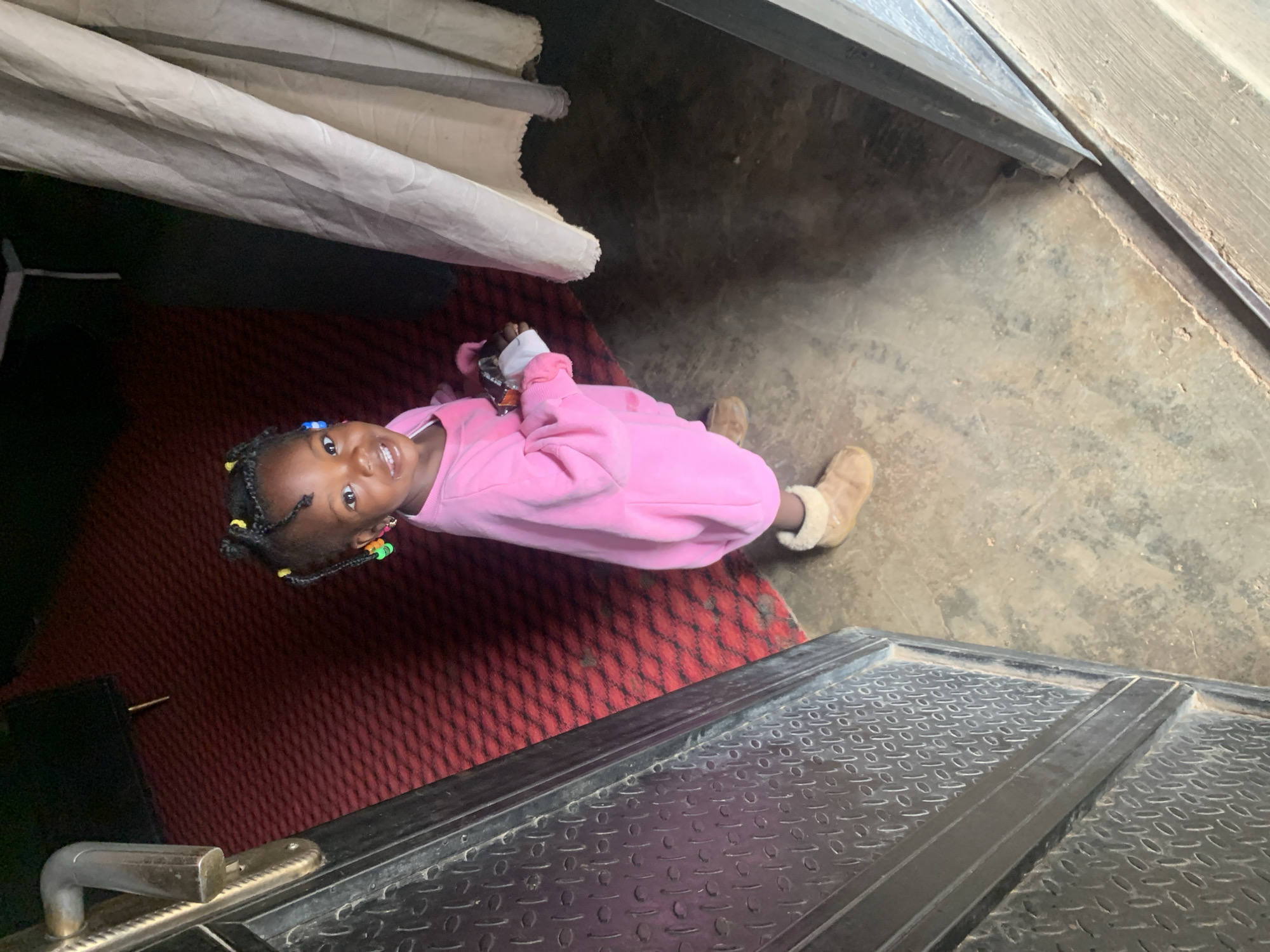 A young child, appearing to be under five years old, stands near an open doorway. The child has dark skin, braided hair adorned with colorful beads, and is wearing a bright pink sweatshirt and tan boots. The child is holding a small, light-colored object in their right hand. The background shows a dark red carpet, a textured metal door frame, and a beige curtain. The floor is a concrete surface with some debris. This scene is located in Jos, Nigeria, likely within a residential or commercial building.